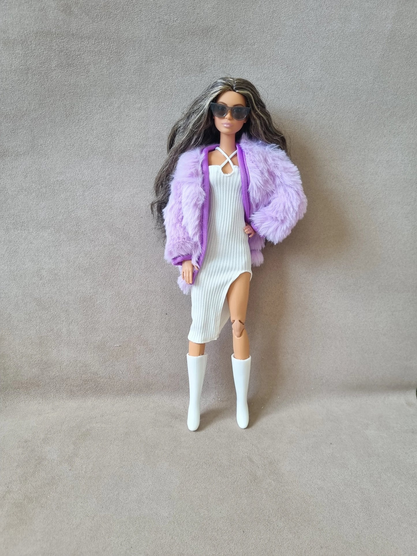 Barbie doll wearing white ribbed dress, lavender faux fur coat, sunglasses, and white knee-high boots against grey background.