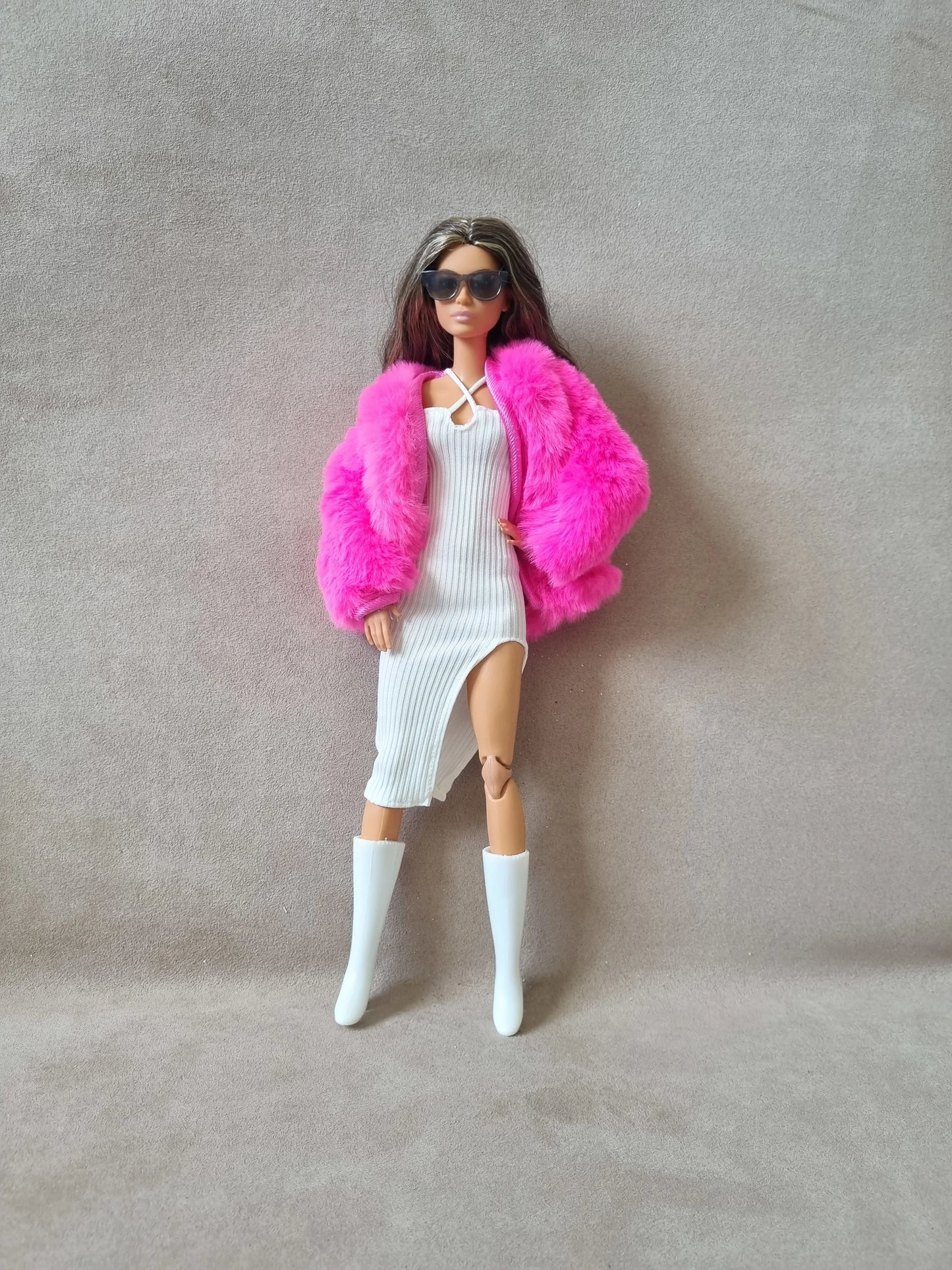 Barbie doll wearing white ribbed dress, hot pink faux fur coat, sunglasses, and white knee-high boots against grey background.