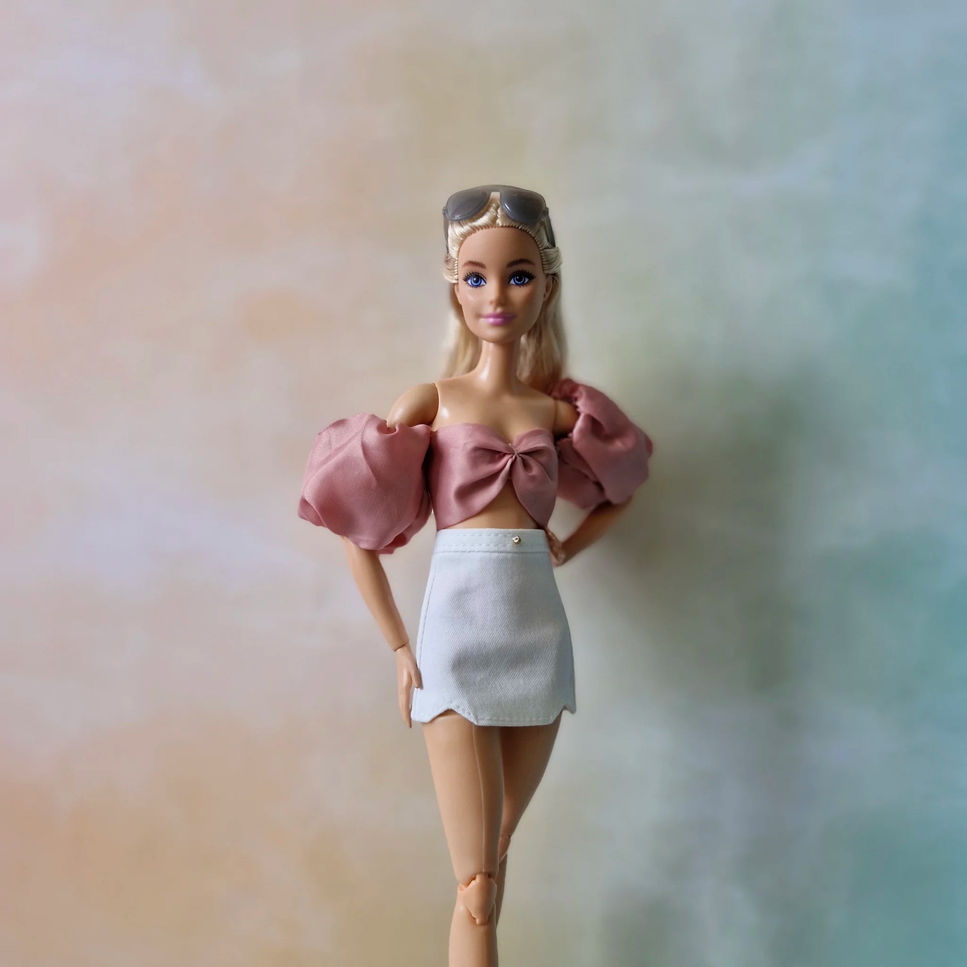 Barbie doll wearing sunglasses on head, dusty rose bow crop top, and white mini skirt, one hand on hip.