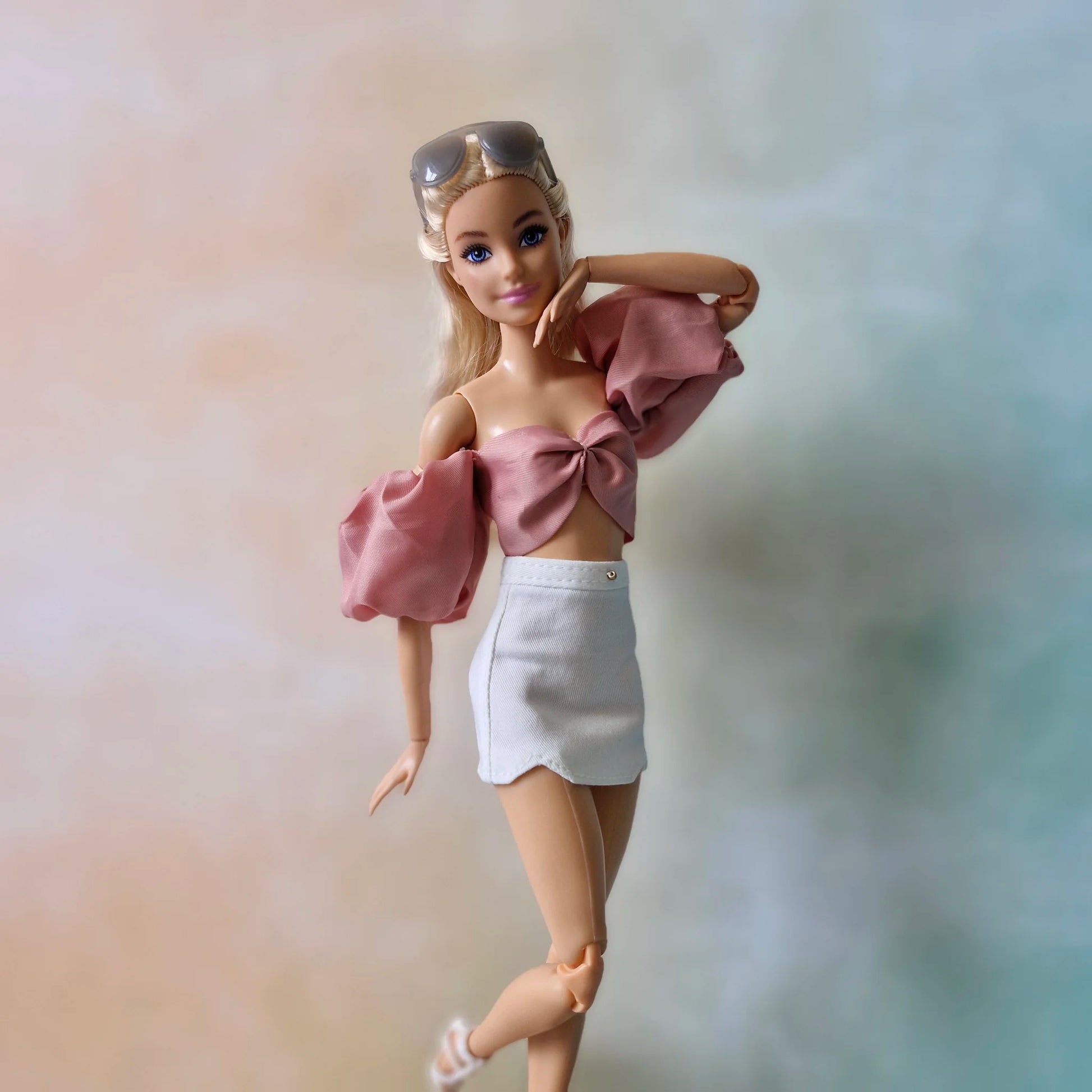 Blonde Barbie doll posing in dusty rose puff sleeve bow crop top and white mini skirt with white heels.