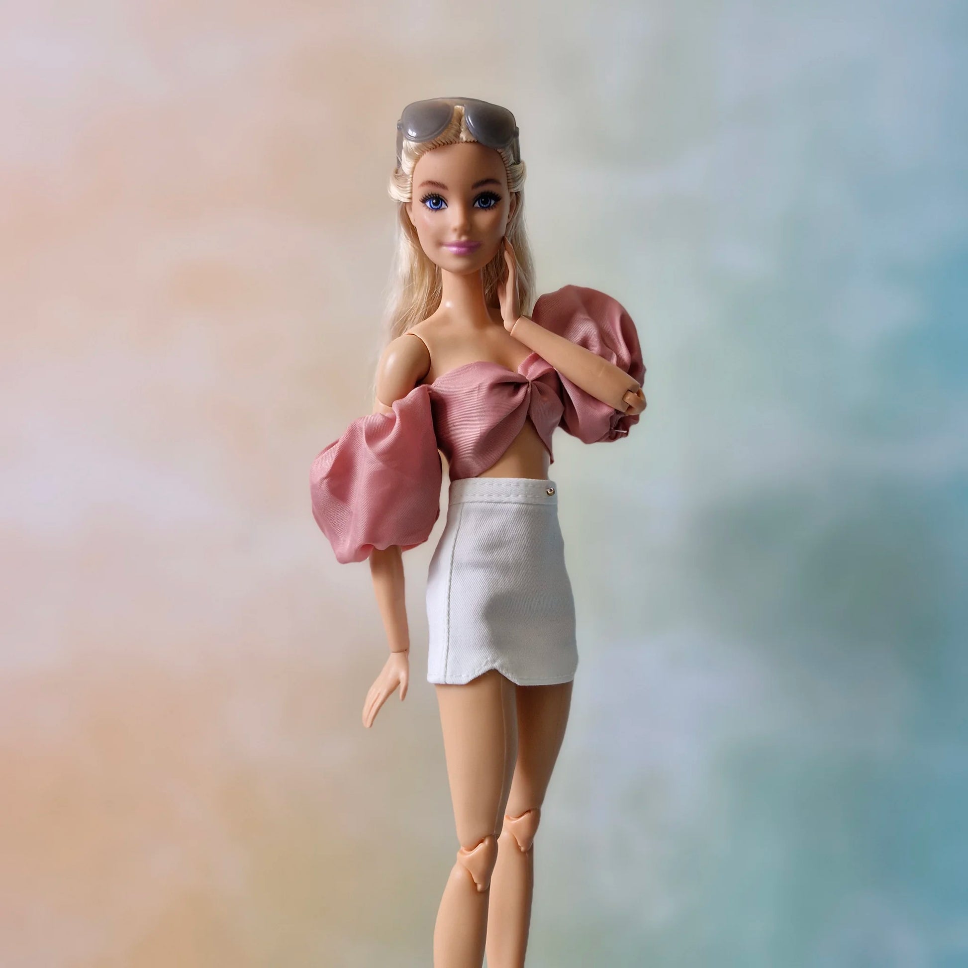 Barbie doll with sunglasses on head wearing dusty rose bow top and white mini skirt, hand near chin.