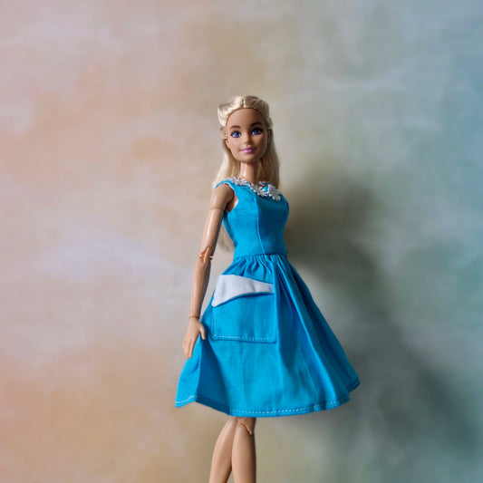 Blonde Barbie doll wearing a blue fit-and-flare dress featuring white applique trim and skirt pockets.