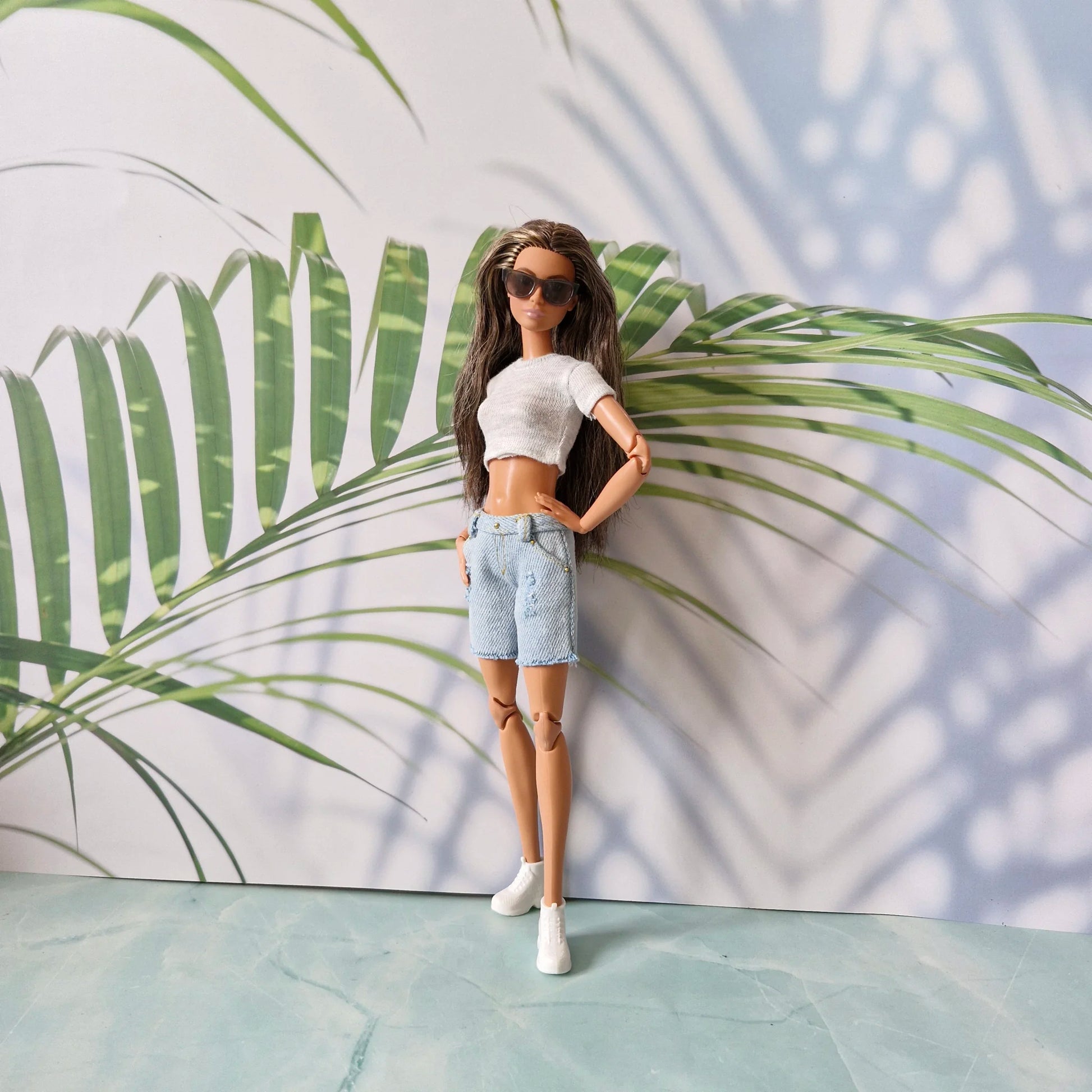 Full view of Barbie doll in sunglasses, grey knit crop top, distressed denim shorts, and white sneakers against tropical leaf background.