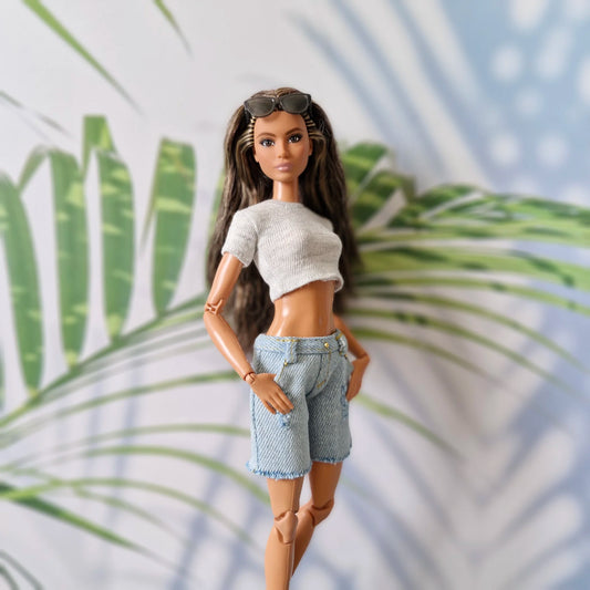 Close-up of Barbie doll wearing grey knit crop top, light wash denim shorts with gold studs, and sunglasses on head against tropical leaf background.