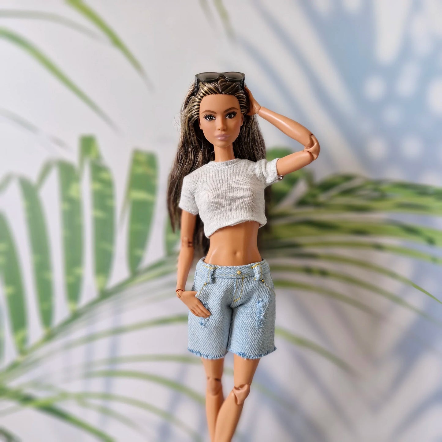 Barbie doll models summer outfit: grey knit crop top, distressed denim shorts, sunglasses on head, hand touching hair, tropical leaf background.