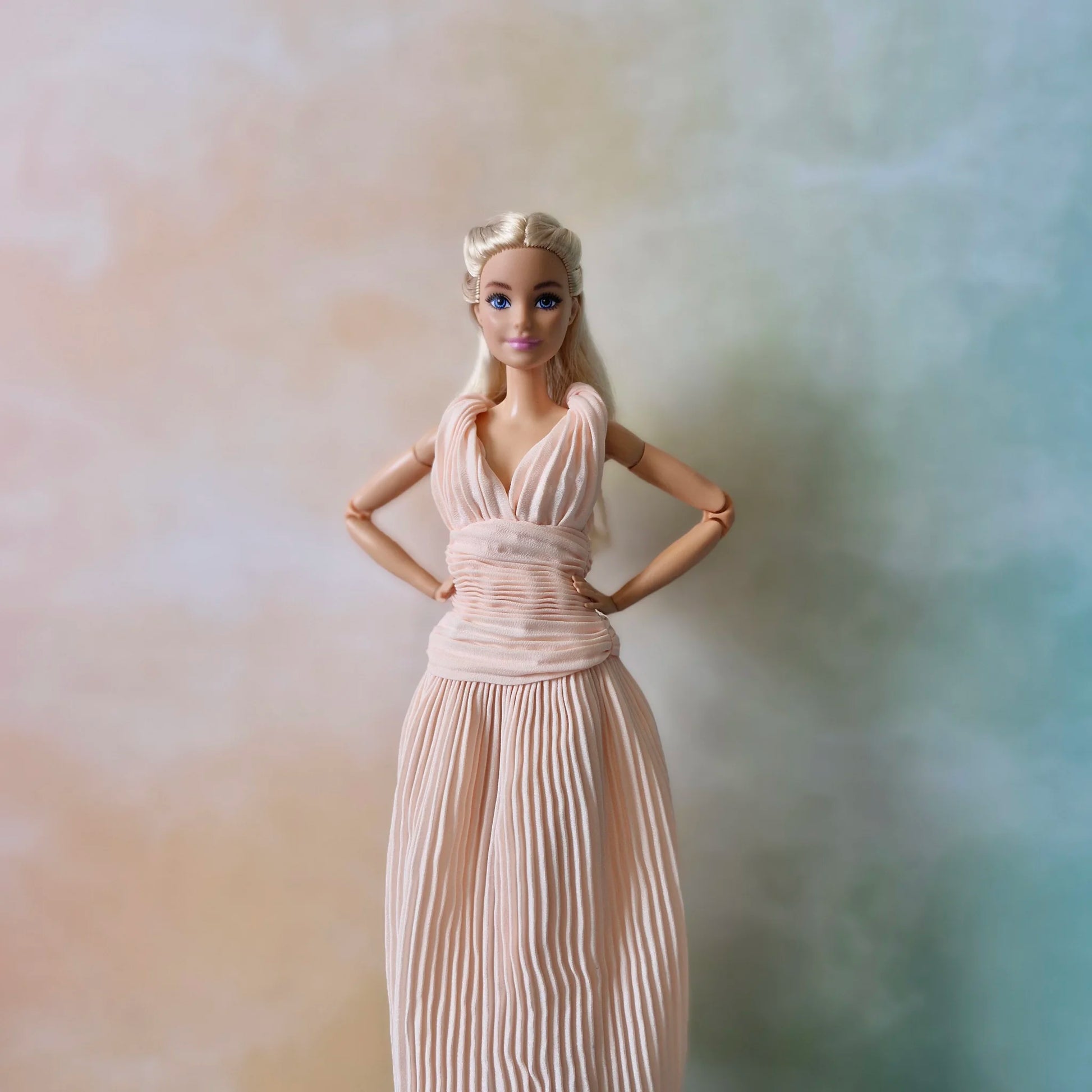 Barbie doll standing confidently with hands on hips, showcasing the fit of the blush pink pleated Grecian gown.