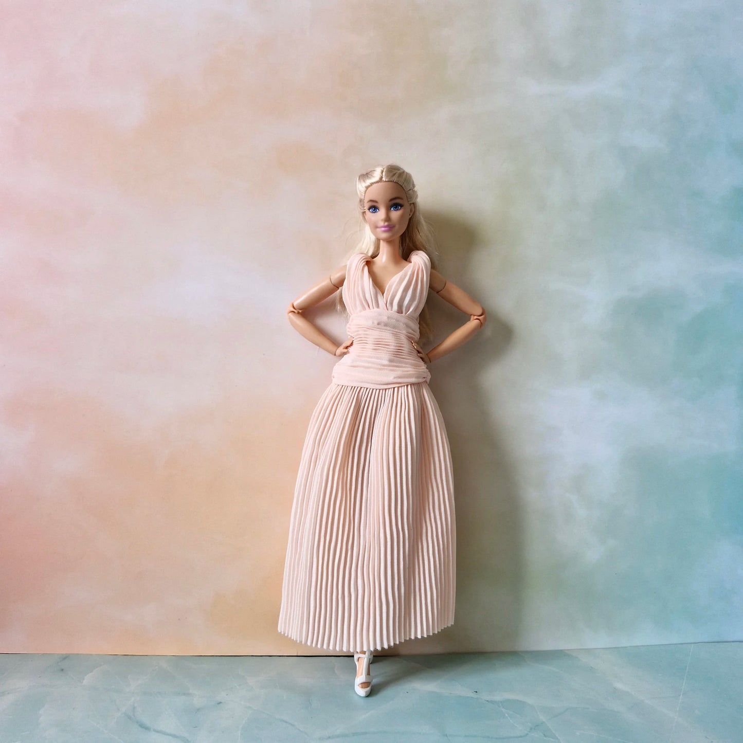 Full length view of Barbie doll standing in blush pink pleated Grecian dress and white heels against pastel background.