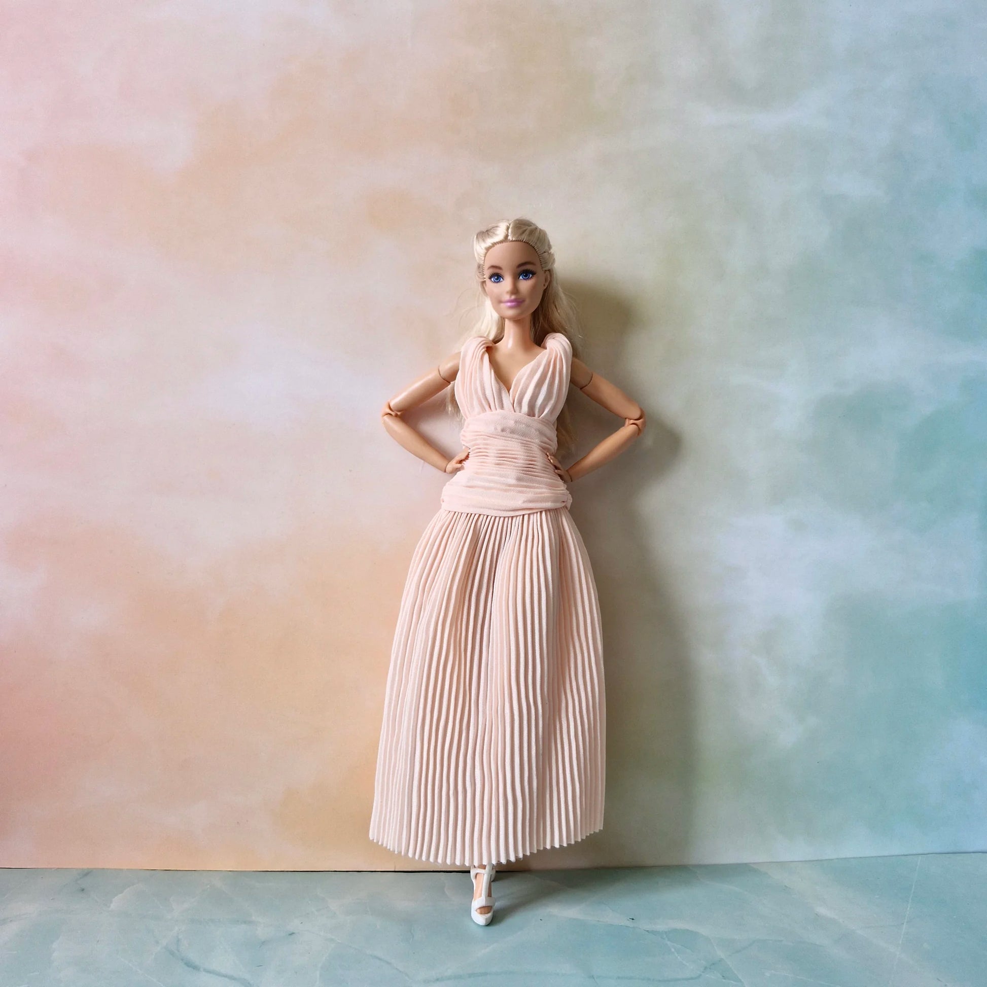 Full length view of Barbie doll standing in blush pink pleated Grecian dress and white heels against pastel background.