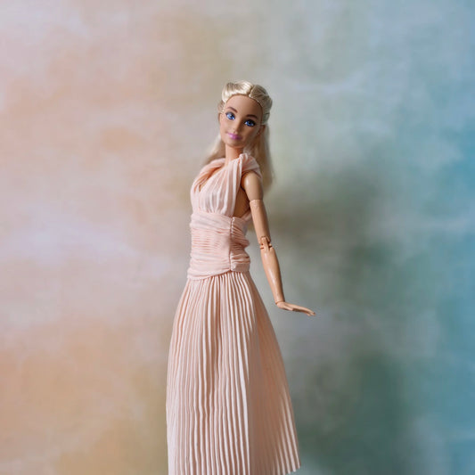 Side profile of blonde Barbie doll modeling blush pink pleated maxi dress, showing draped bodice and flowing skirt.