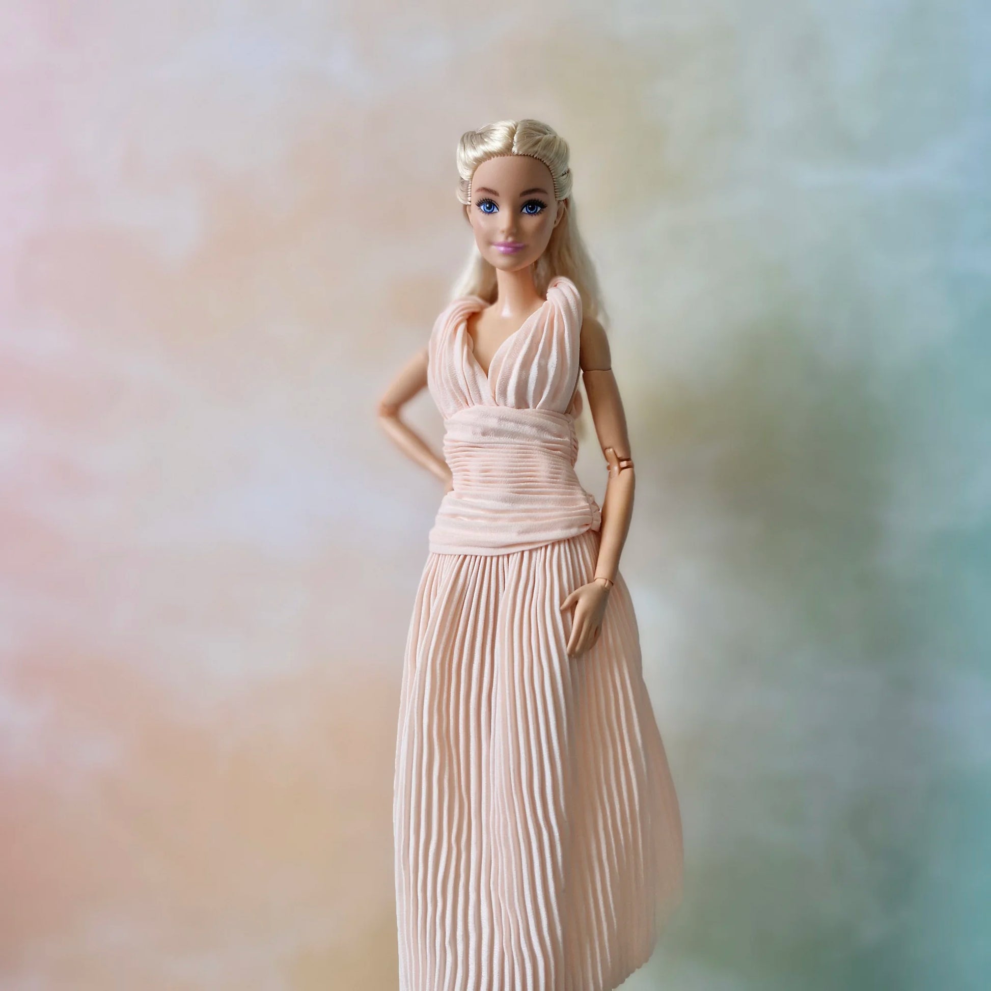 Blonde Barbie doll posing, hand on hip, in elegant blush pink pleated Grecian gown with ruched bodice.