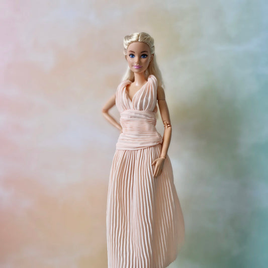 Blonde Barbie doll posing, hand on hip, in elegant blush pink pleated Grecian gown with ruched bodice.