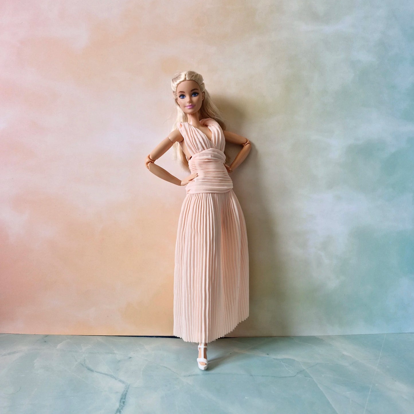 Barbie doll leaning against wall, posing in elegant full-length blush pink pleated dress and white shoes.