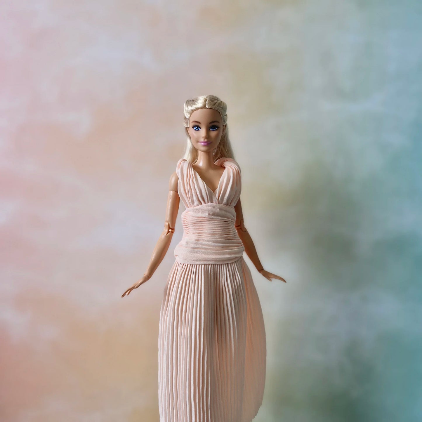 Front view of blonde Barbie doll wearing sophisticated blush pink pleated maxi dress with draped bodice detail.