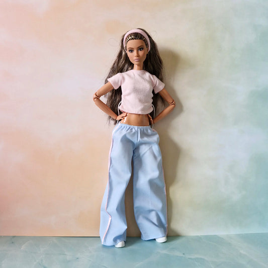 Barbie doll with long brown hair models athleisure outfit: pink drawstring crop top and blue wide-leg track pants.