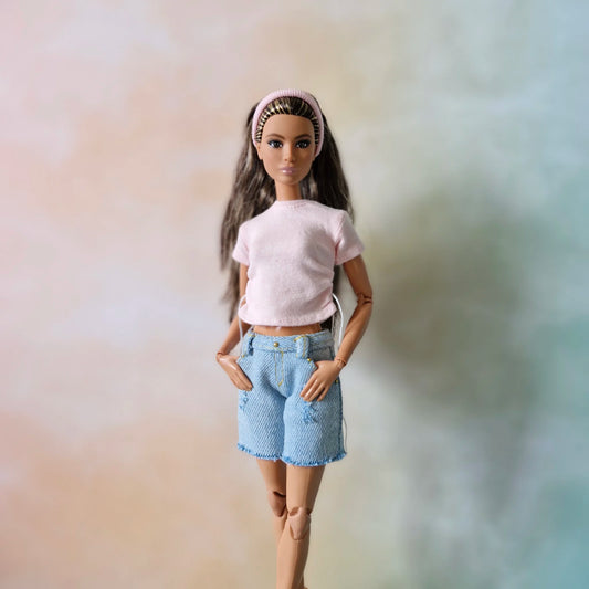 Brunette Barbie doll wearing pink drawstring top, light wash distressed denim shorts, and pink headband, hands in pockets.