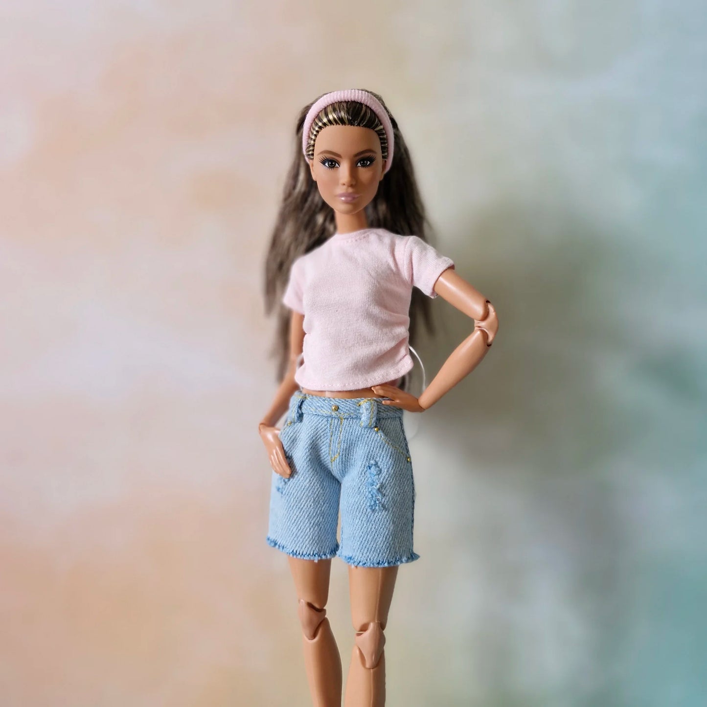 Brunette Barbie doll poses in pink drawstring t-shirt, light blue distressed denim shorts with frayed hem, and pink headband.