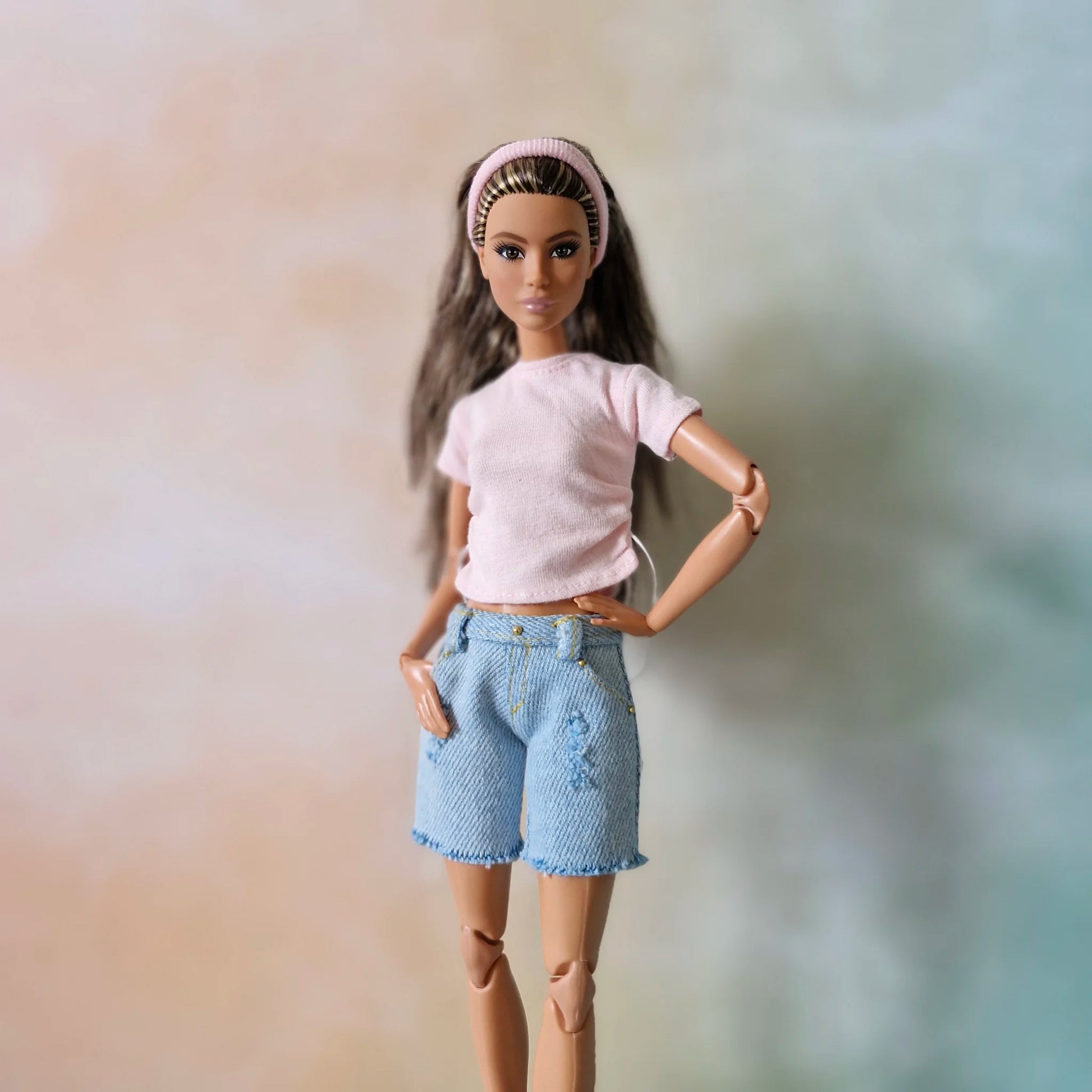 Brunette Barbie doll poses in pink drawstring t-shirt, light blue distressed denim shorts with frayed hem, and pink headband.