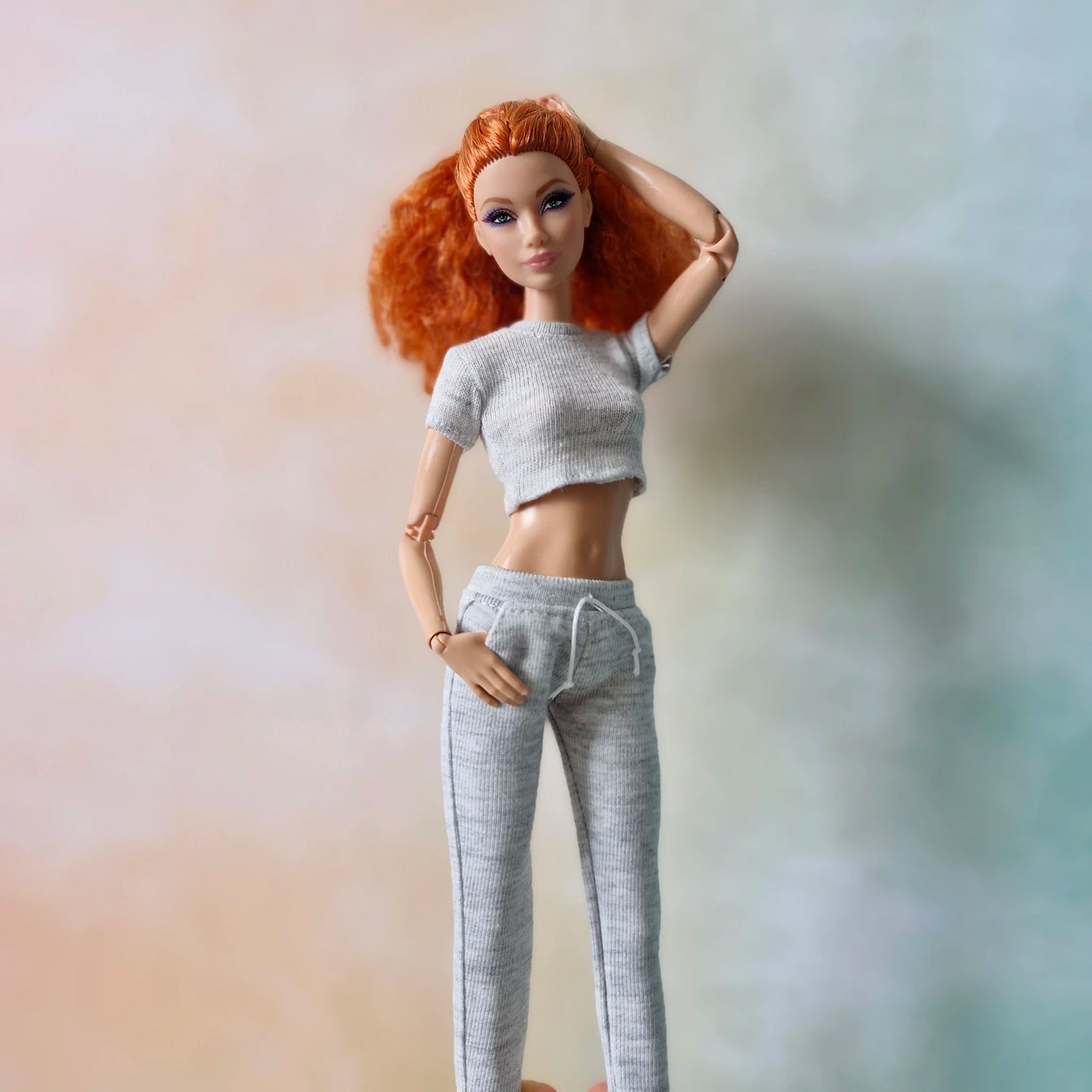 Redhead fashion doll wearing light grey knit crop top and matching joggers, hand touching hair.