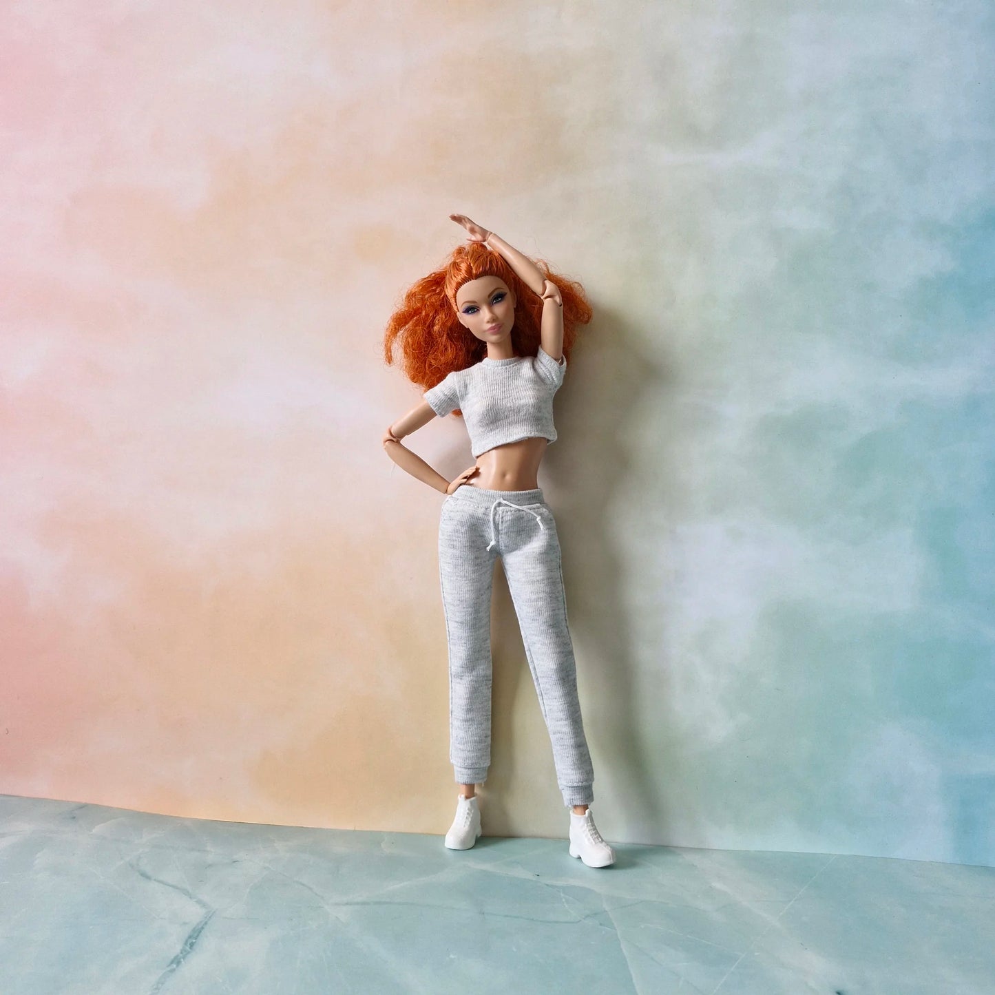 Full view of redhead Barbie doll in grey knit athleisure outfit: crop top, joggers, and white sneakers, posing against pastel background.