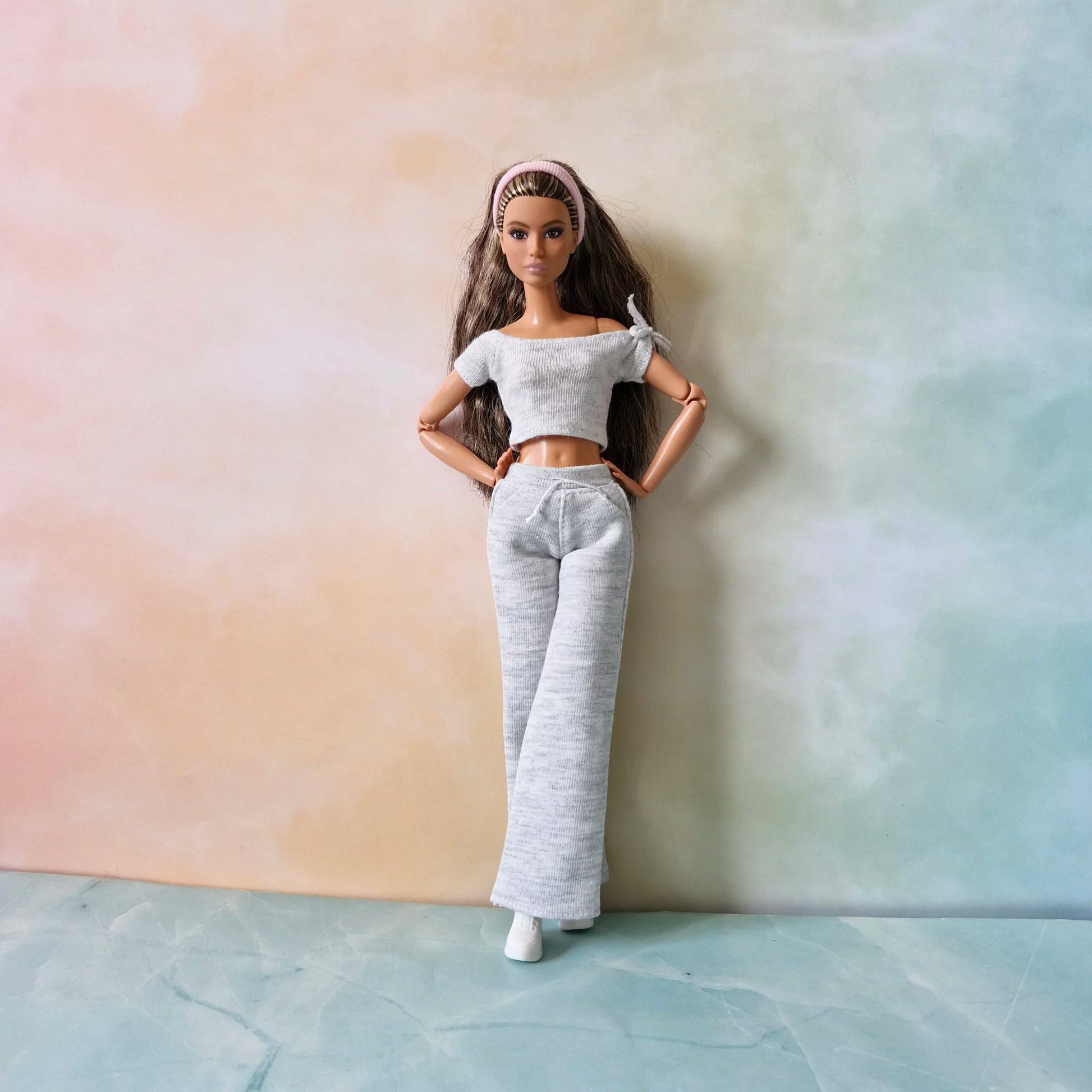 Barbie doll with pink headband models grey knit loungewear: off-shoulder tie-sleeve top and wide-leg pants, hands on hips.