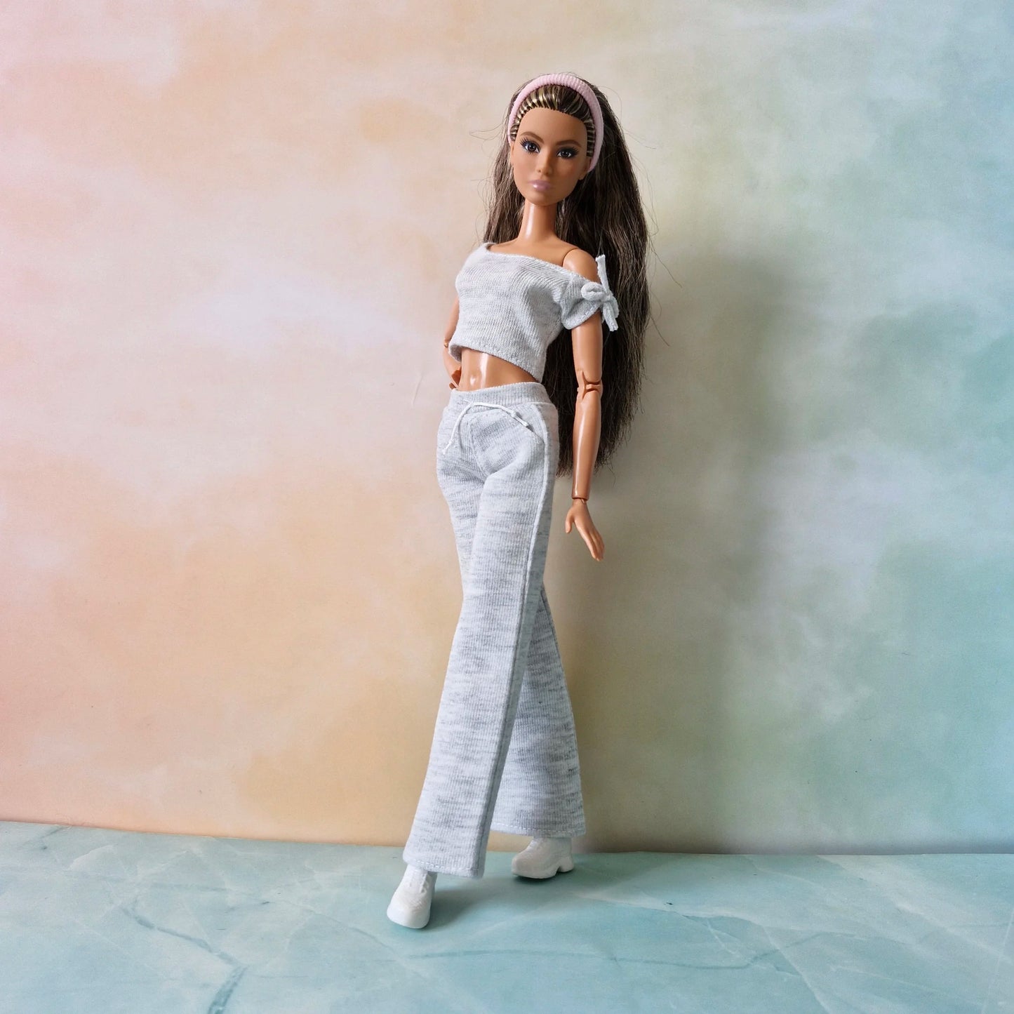 Brunette Barbie doll models grey knit set: off-shoulder crop top, wide-leg pants, and pink headband, angled pose.