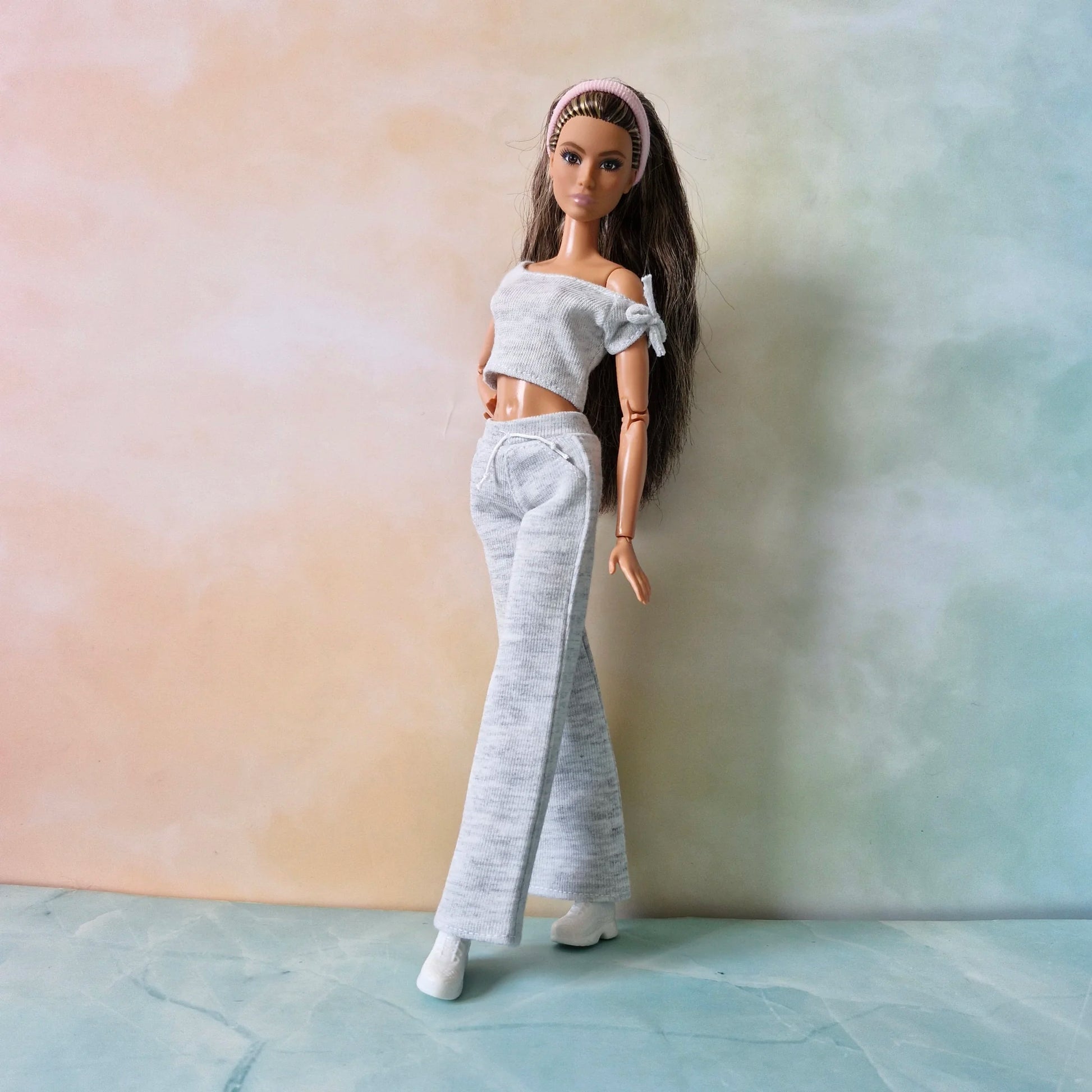 Brunette Barbie doll models grey knit set: off-shoulder crop top, wide-leg pants, and pink headband, angled pose.