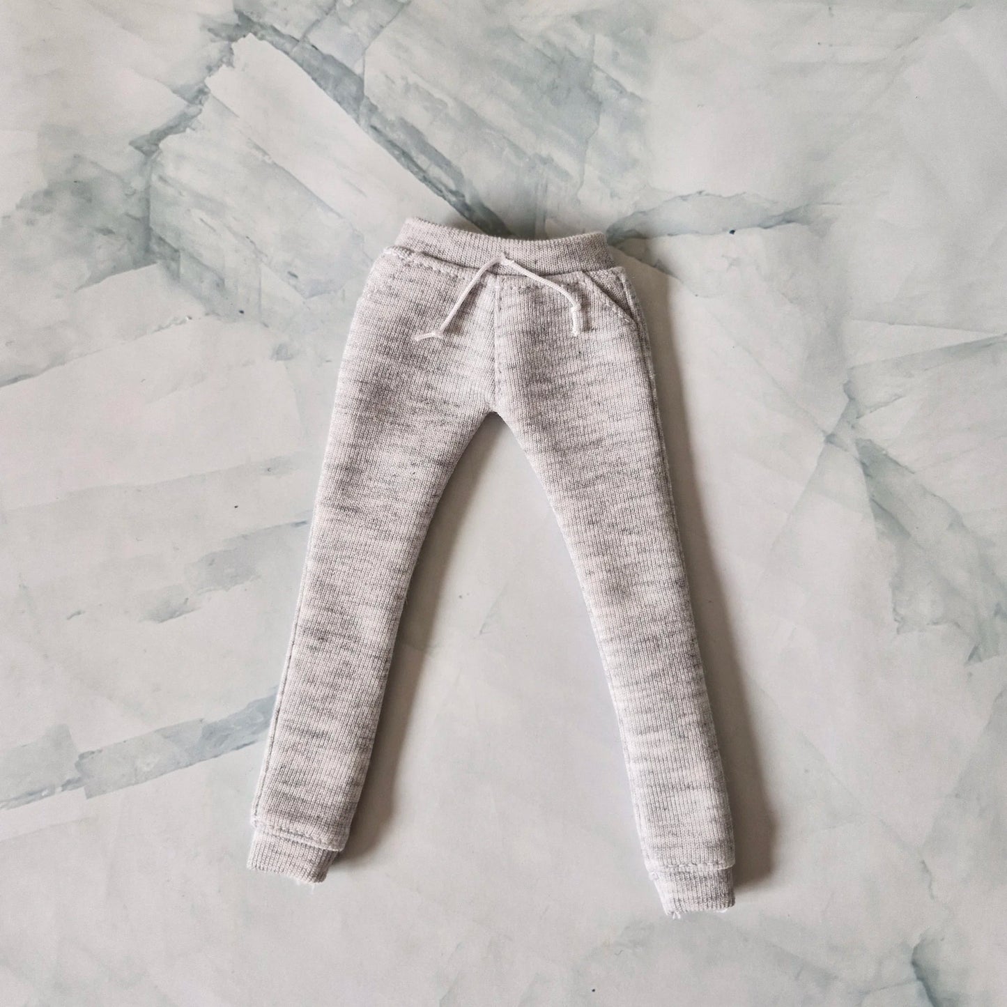 Light grey heather knit joggers for Barbie doll with drawstring waist detail, laid flat on marble background.