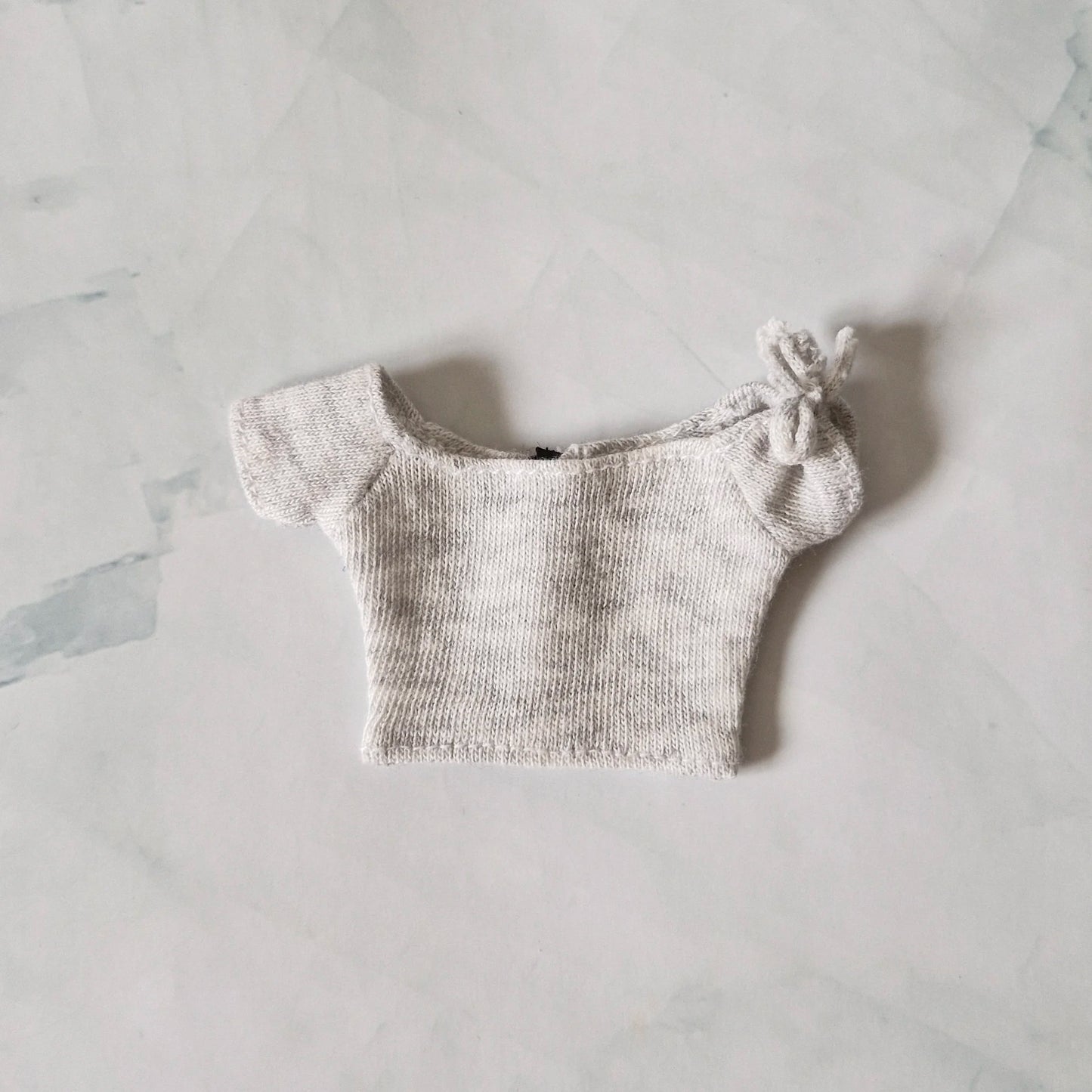 Light grey heather knit off-the-shoulder crop top for Barbie doll with tie detail on sleeve, laid flat on marble background.