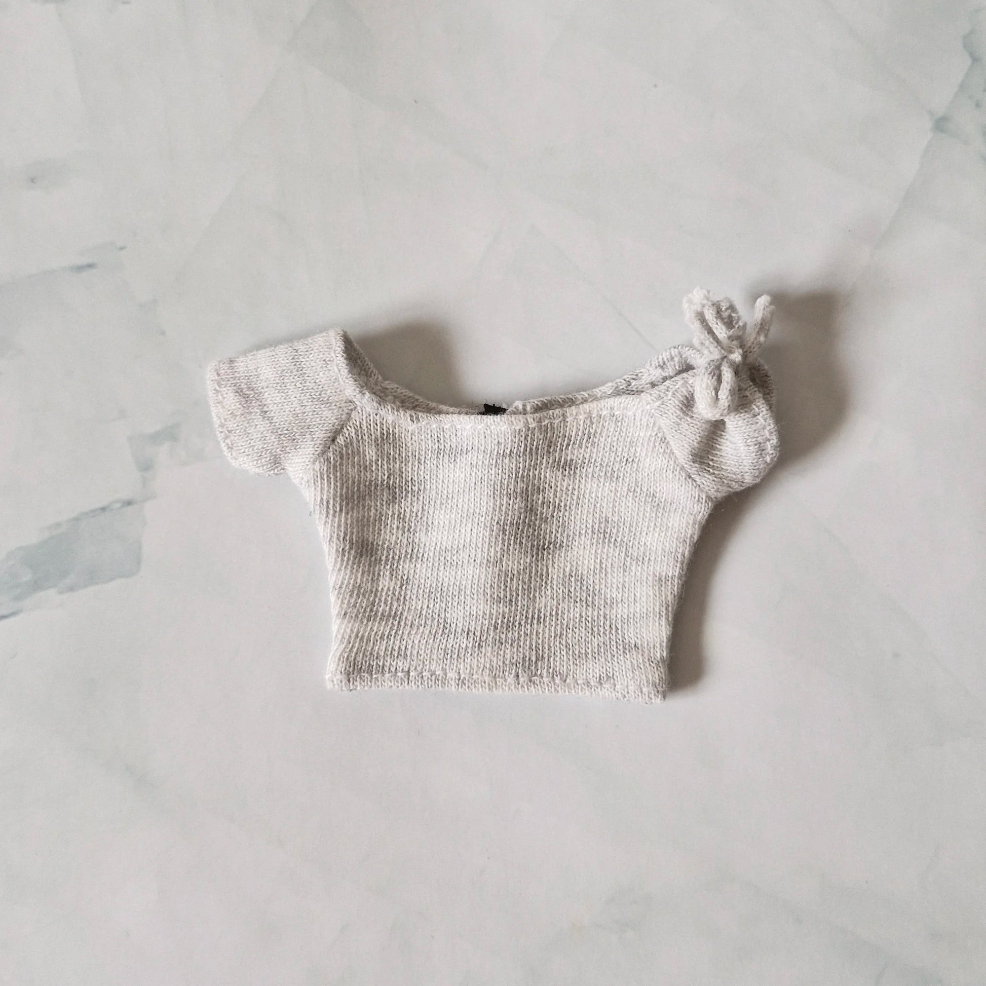 Light grey heather knit off-the-shoulder crop top for Barbie doll with tie detail on sleeve, laid flat on marble background.