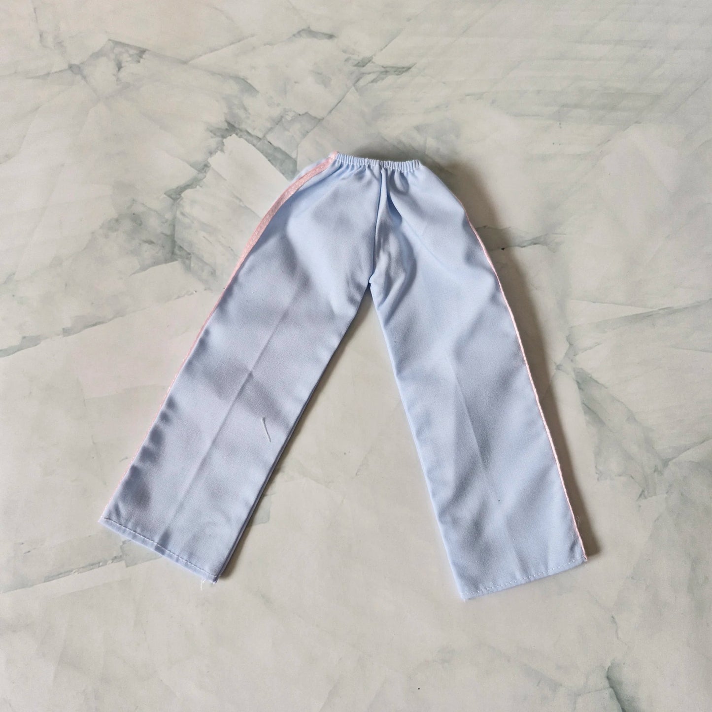 Light blue wide-leg track pants for Barbie doll with pink side stripes, laid flat on marble background.