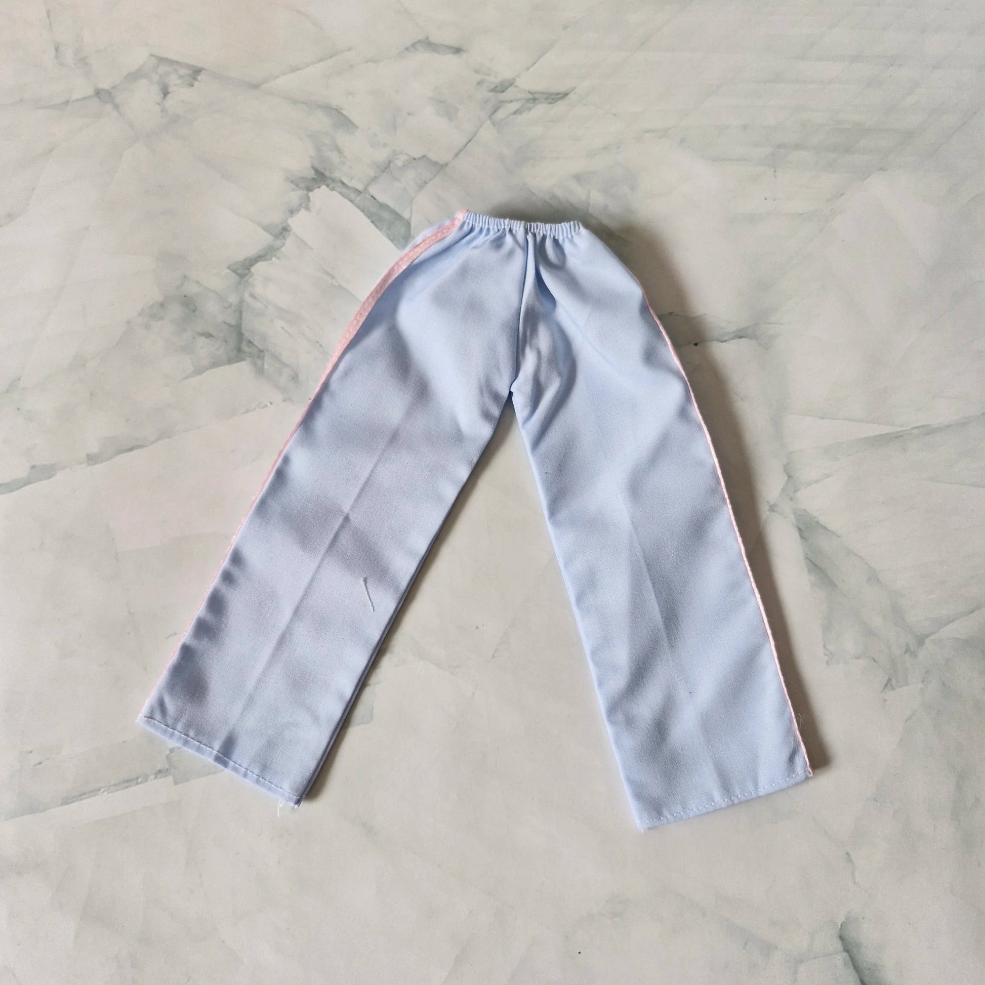 Light blue wide-leg track pants for Barbie doll with pink side stripes, laid flat on marble background.