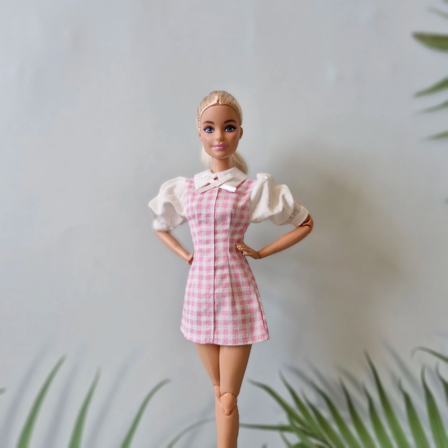 Blonde Barbie doll in a pink and white gingham pinafore dress over a white puff-sleeve blouse, posing with hands on hips.