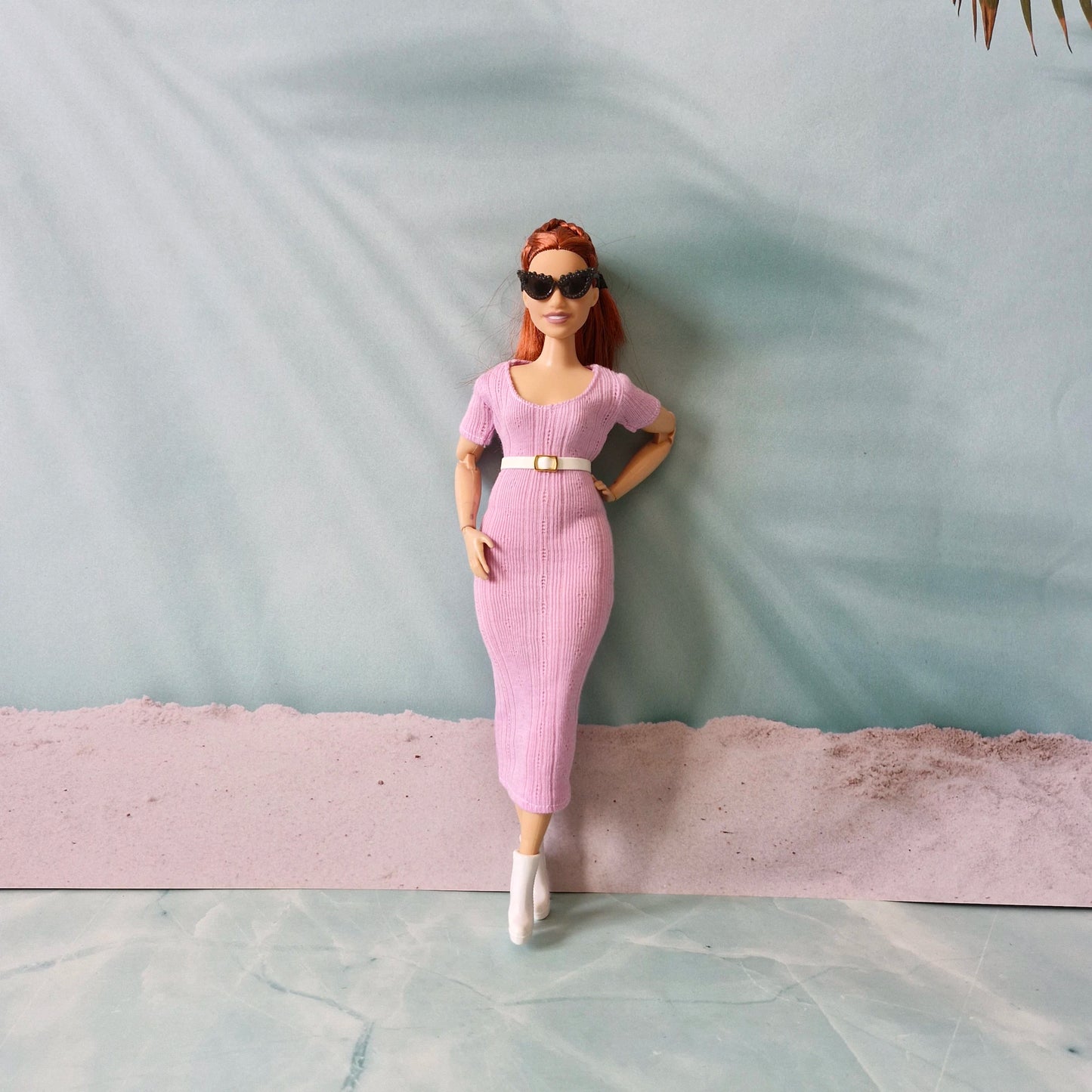 Barbie doll wearing pink ribbed midi dress with a white belt and white boots, leaning against a wall.