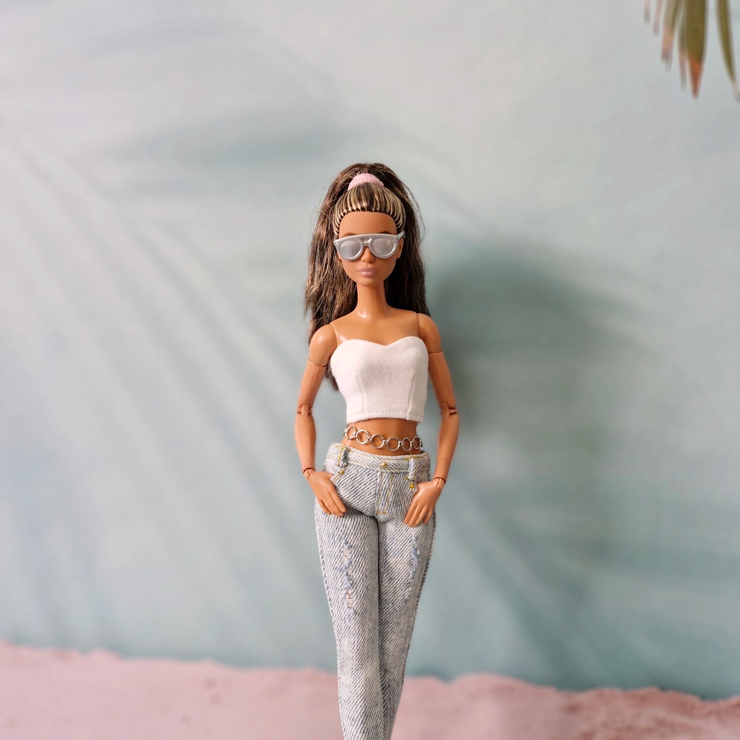 Brunette Barbie doll wearing sunglasses, a white tube top, and distressed jeans with a chain belt detail.