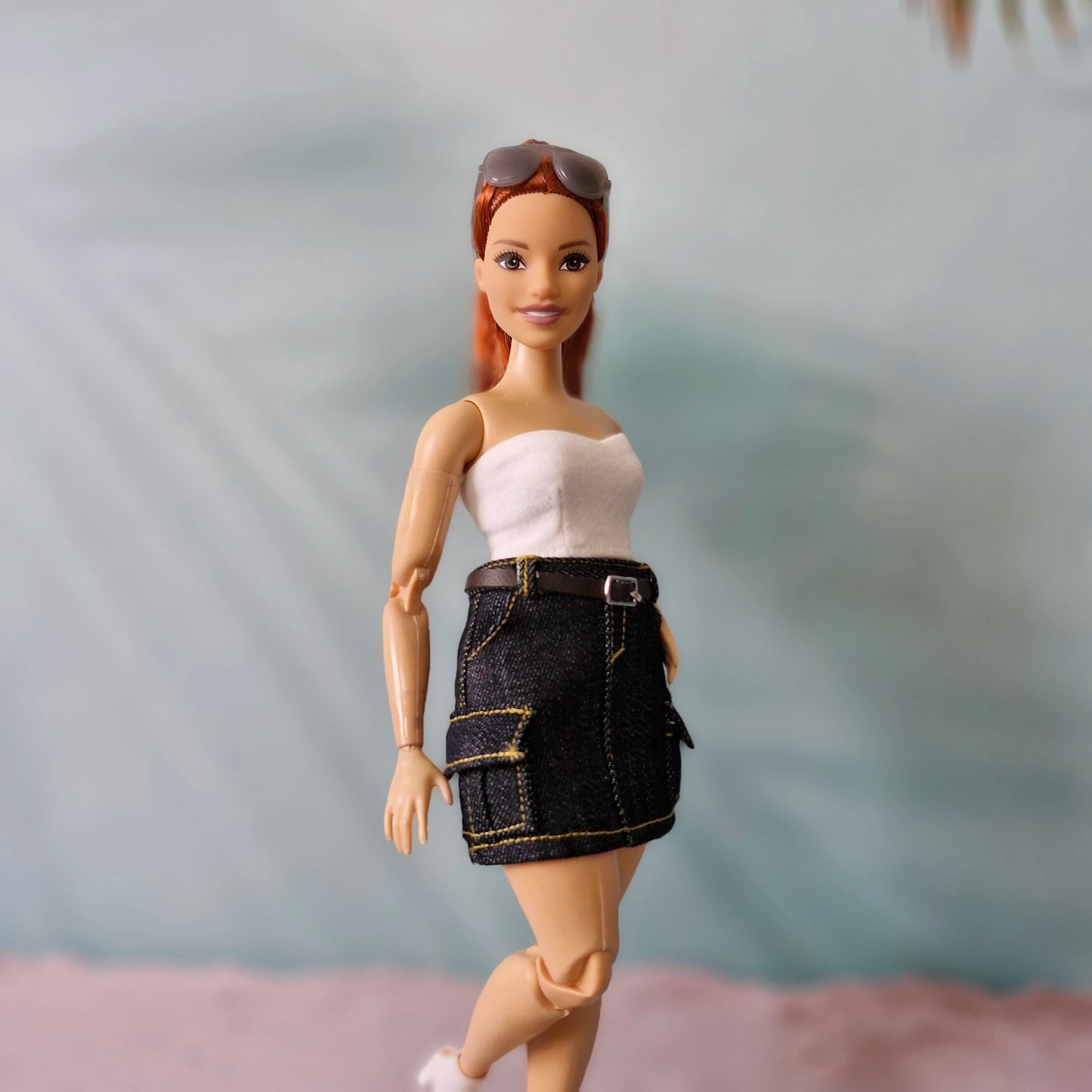 Barbie doll wearing a white top and black denim skirt with sunglasses on head against a blurred background