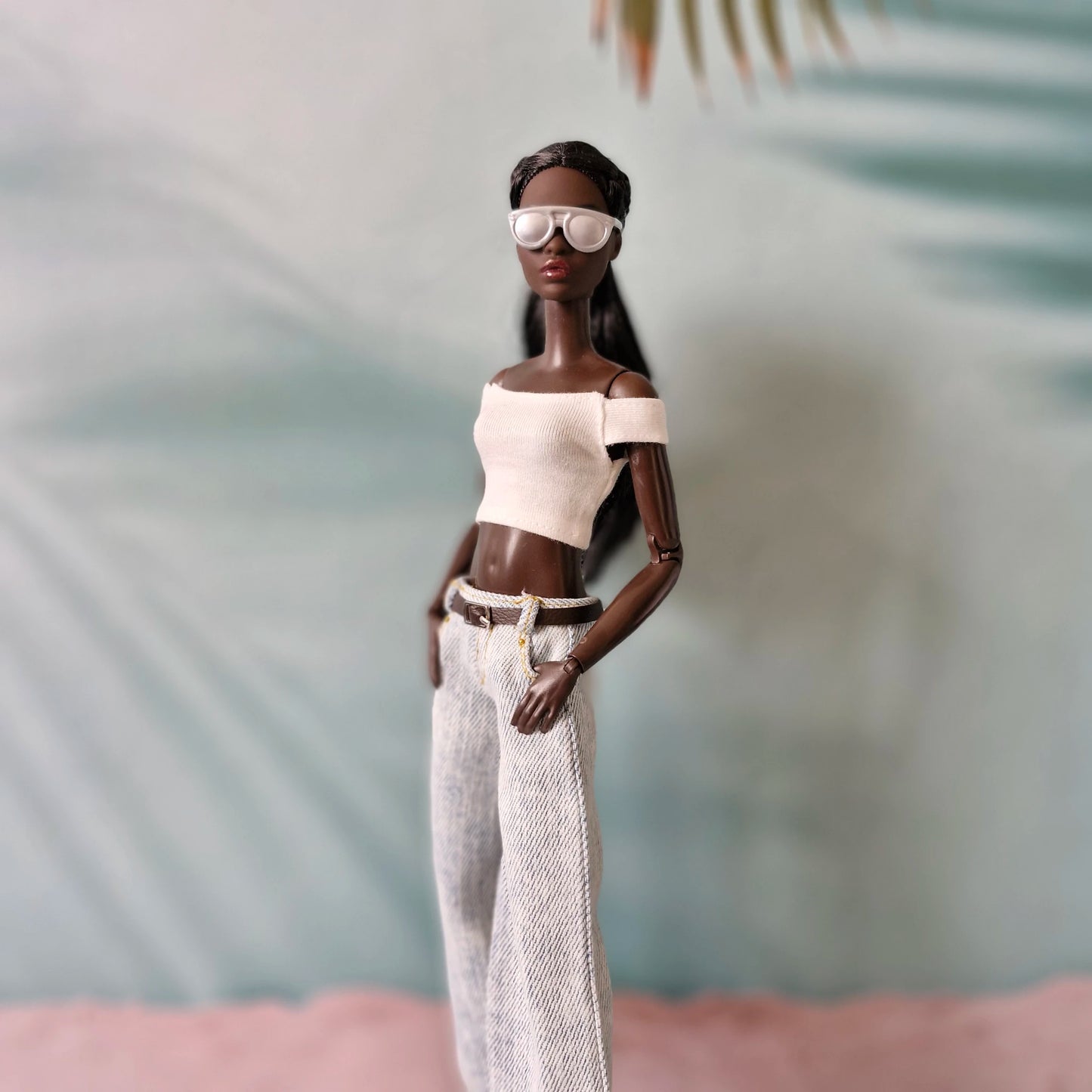 Doll wearing a white top and pants with sunglasses against a blurred natural background