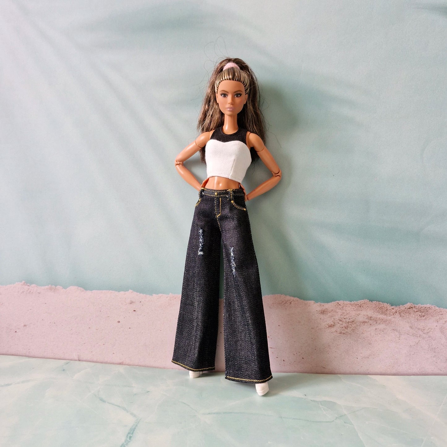 Barbie doll wearing a white top and black jeans standing on a textured surface with a light blue background.