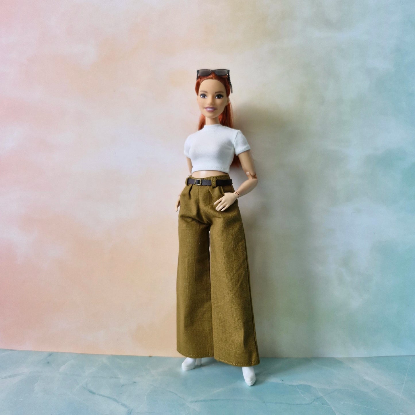 Barbie doll with red hair wearing a white top and brown pants against a gradient background