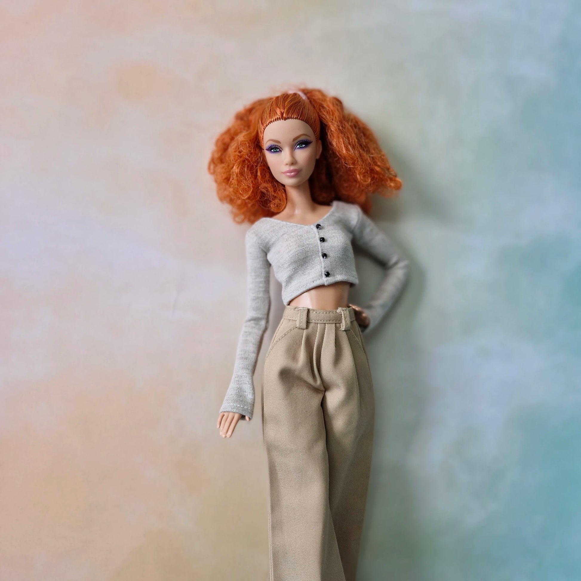 Barbie doll wearing a gray sweater and beige pants against a gradient background