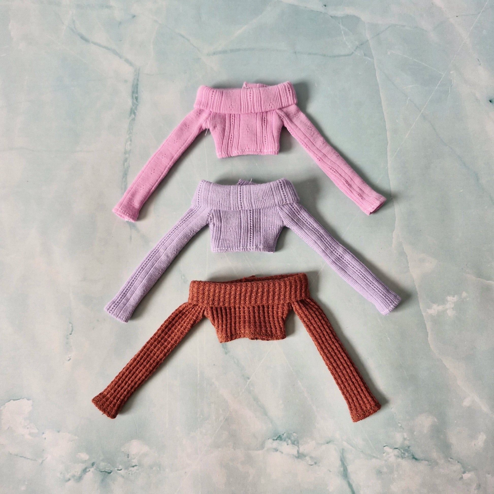 Three knitted tops in pink, purple, and brown on a textured gray surface