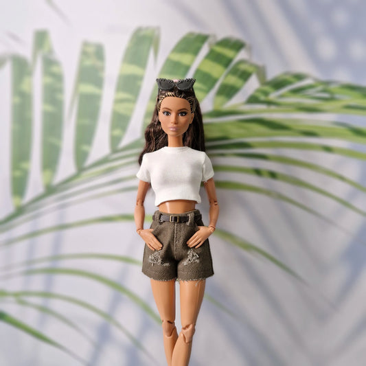 Barbie doll wearing a white top and brown shorts with a palm leaf background