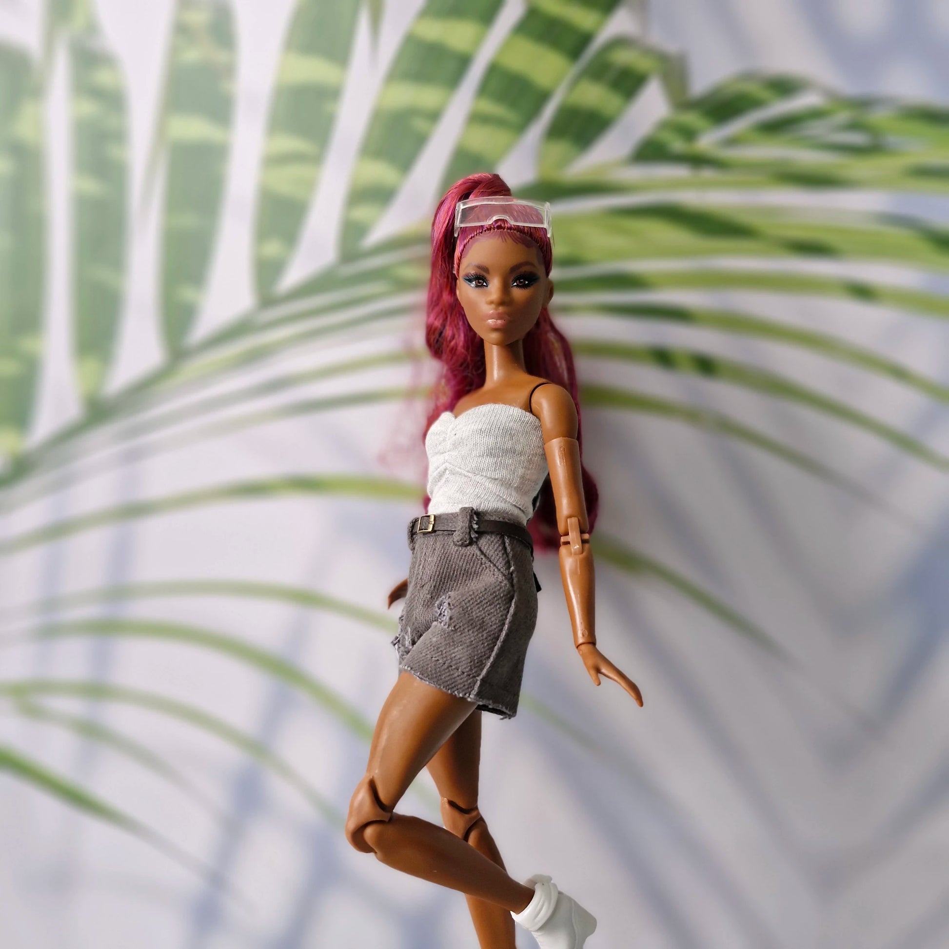 Barbie doll with pink hair and white top in front of palm leaves