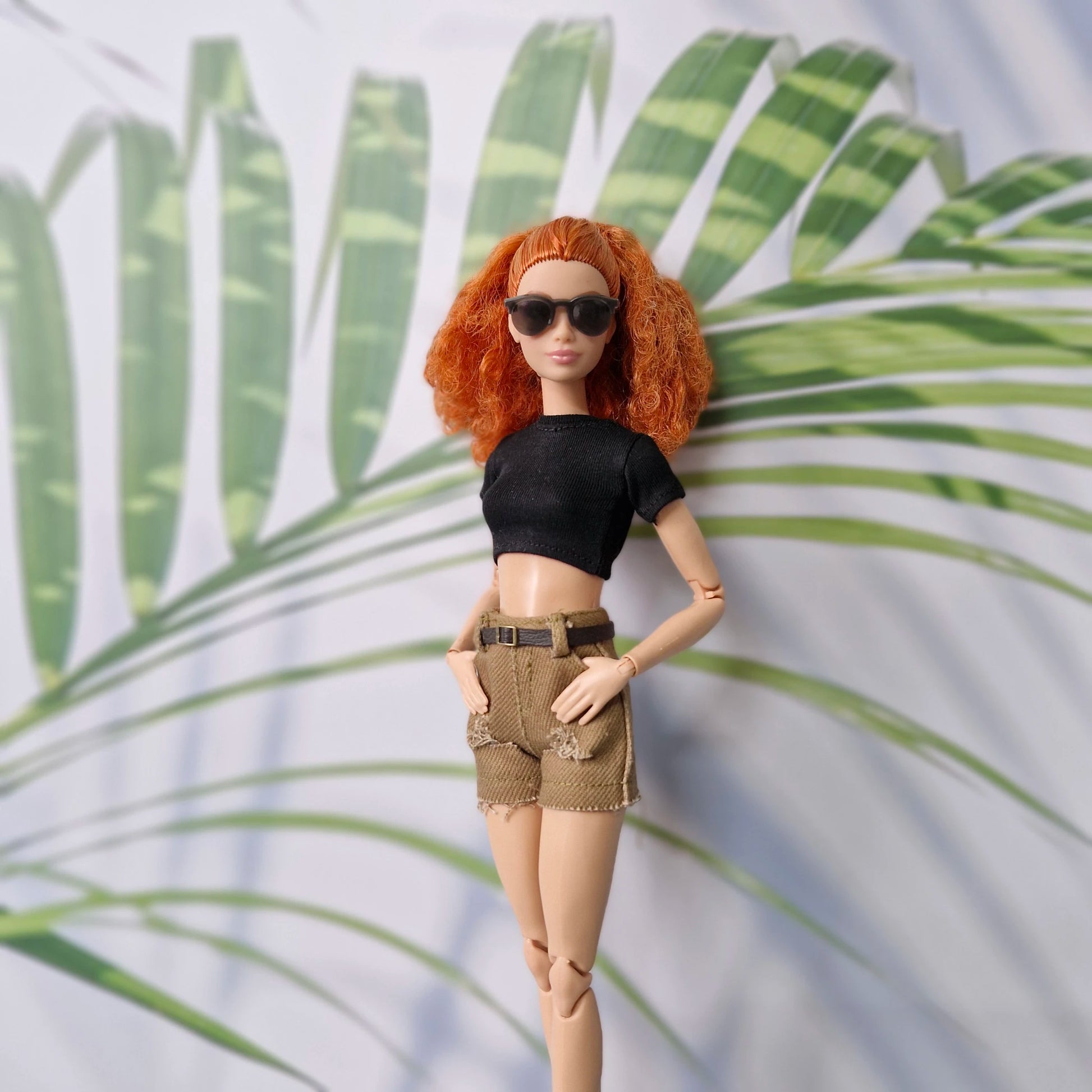 Barbie doll with red hair and sunglasses standing against a leafy background