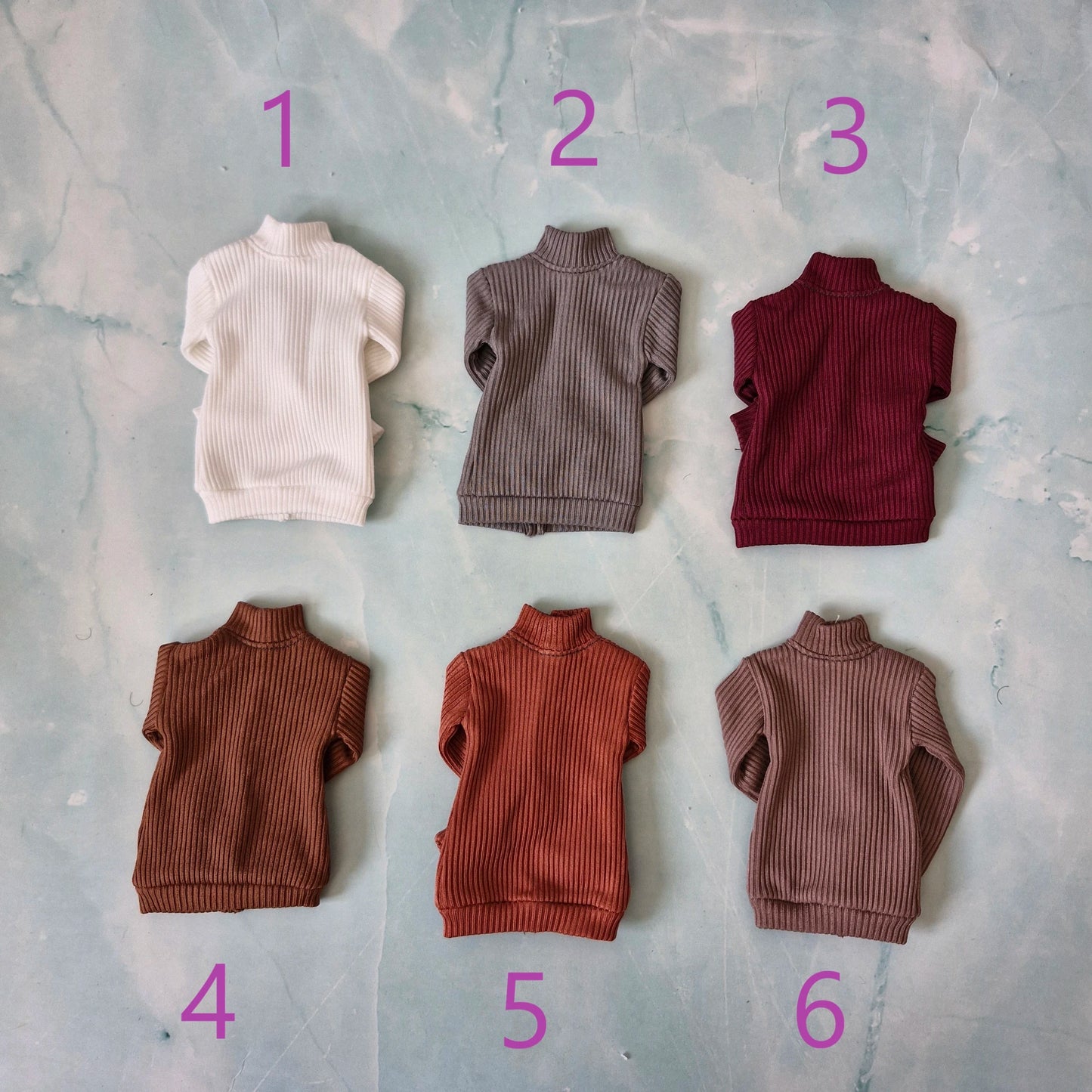 Six miniature ribbed knit turtleneck sweaters for dolls, numbered 1 to 6 in purple text, arranged on a light blue marbled background.