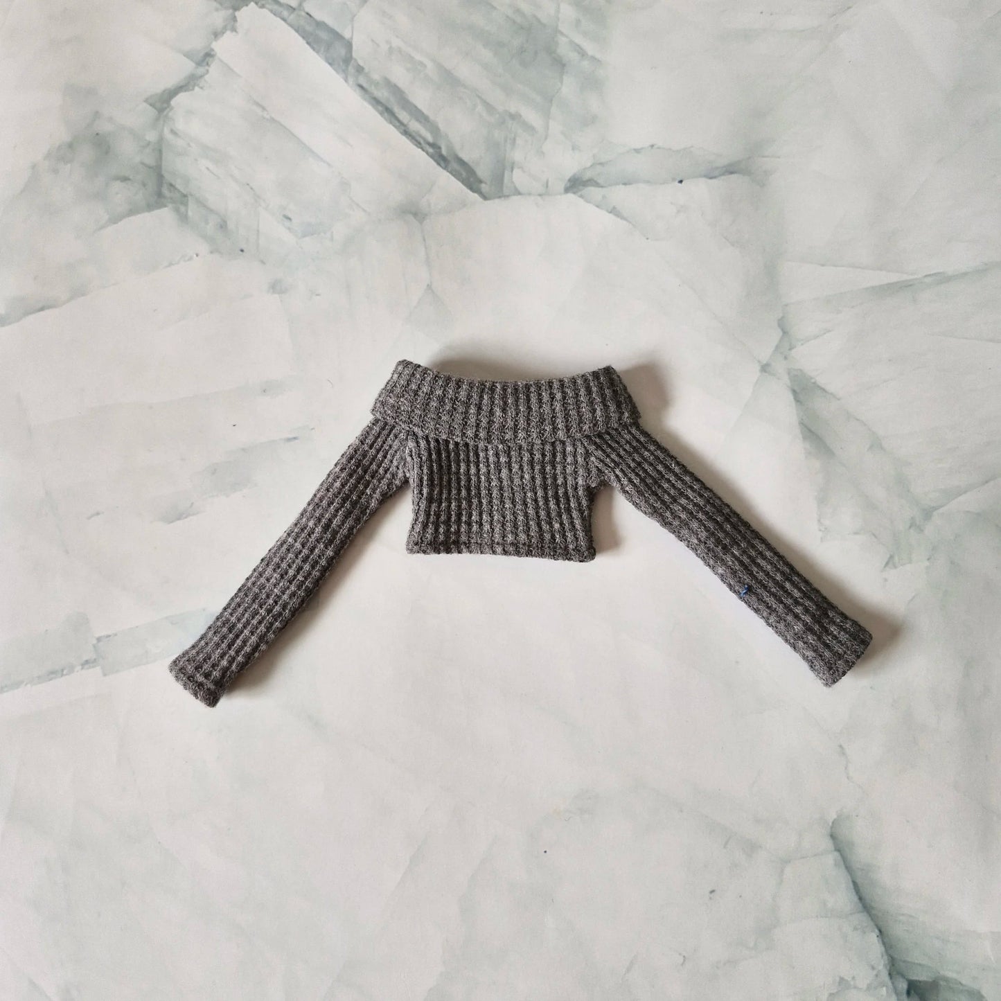 Flat lay of grey ribbed knit off-shoulder long-sleeve crop top sweater for fashion dolls on a marble background.