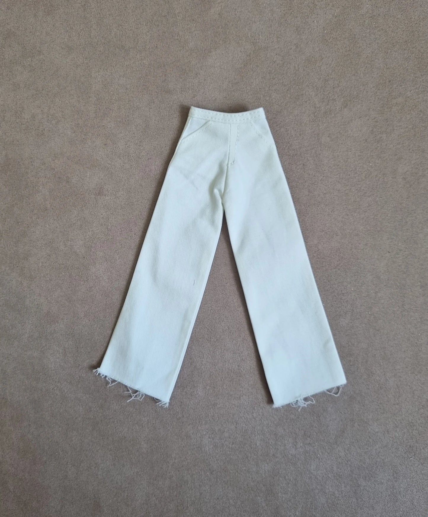 Handmade white wide-leg pants for Barbie dolls, featuring denim-style texture and frayed hems, lying flat on a brown surface
