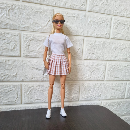 Blonde Barbie doll modeling casual outfit: white t-shirt, pink plaid pleated mini skirt, sunglasses, white bag with chain strap, and white sneakers, standing against white brick wall.