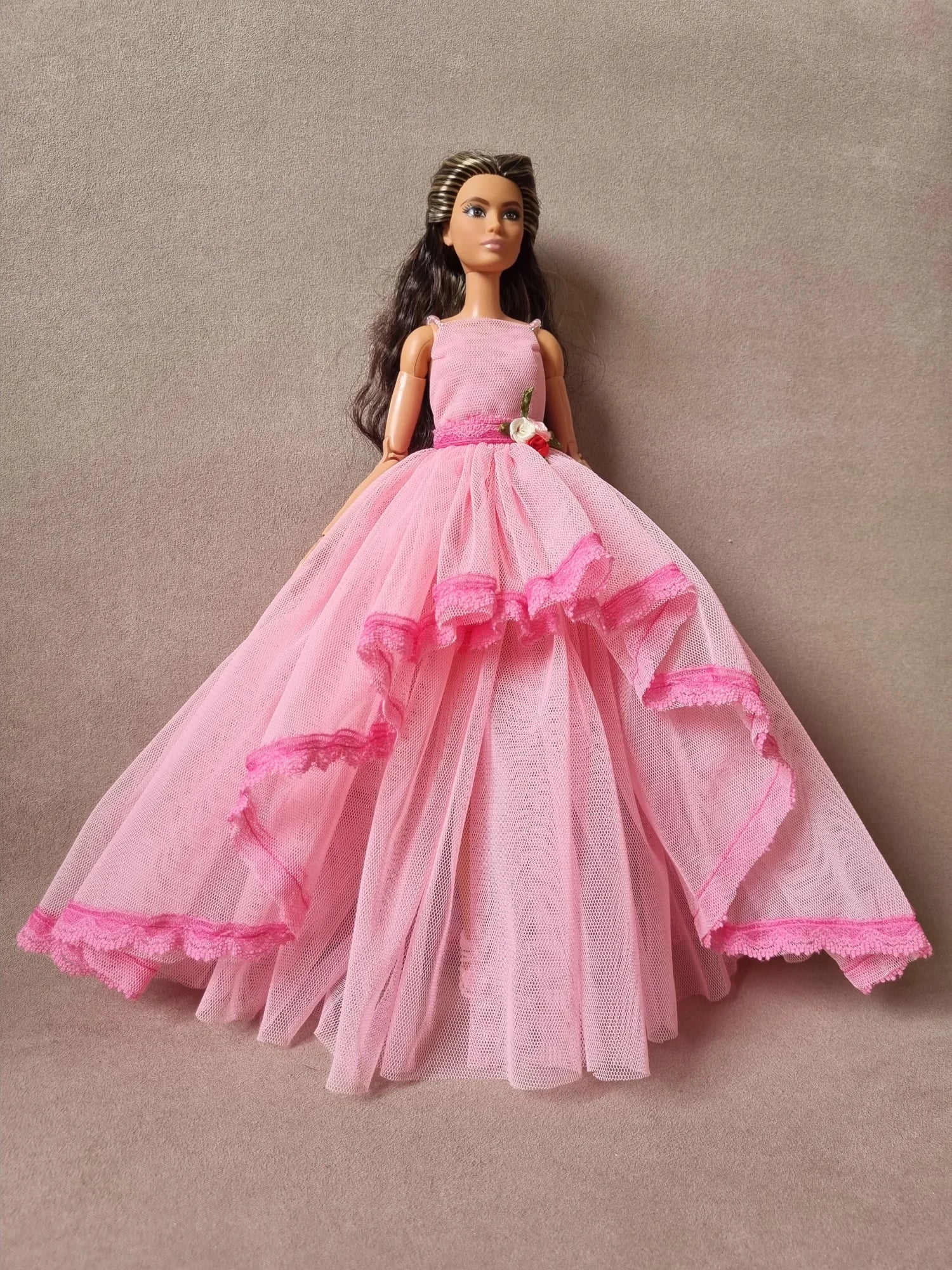 Barbie doll models stunning pink layered tulle ball gown, sleeveless with floral waist detail against beige background.