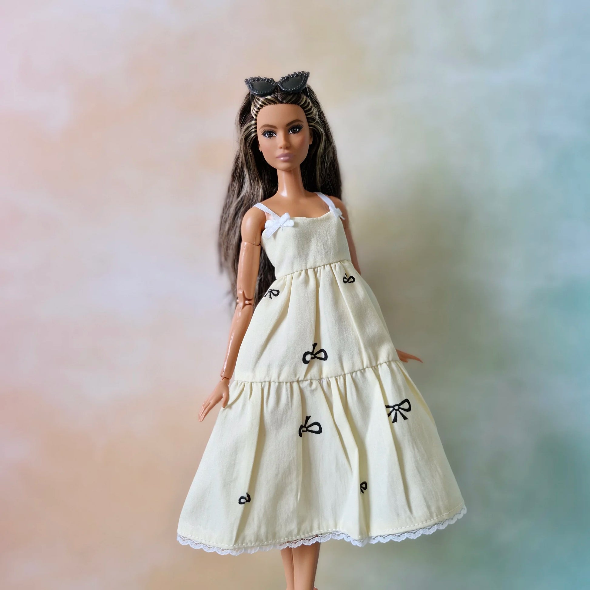 Brunette fashion doll wearing cream tiered sundress with black bow print and white lace hem.