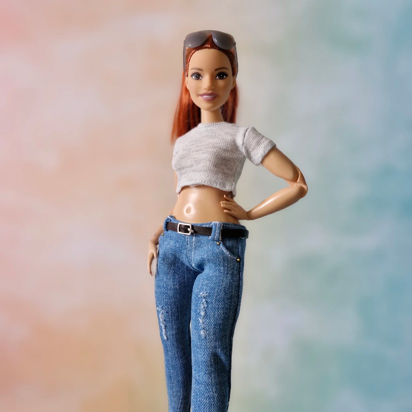 Redhead Barbie doll poses in grey crop top and distressed blue skinny jeans with belt, hand on hip